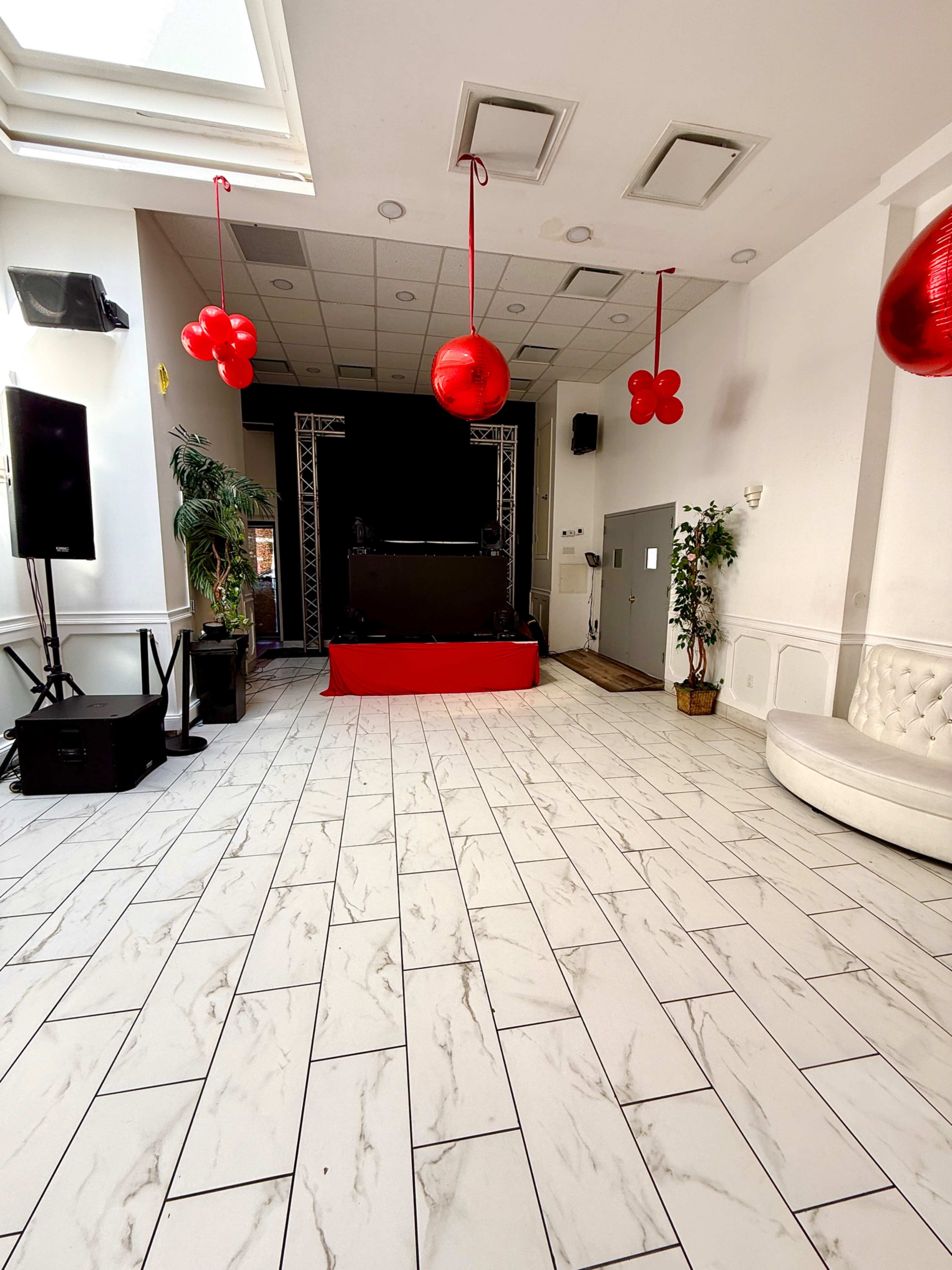 A spacious indoor venue features a raised DJ platform, a large speaker setup, and decorative red balloons hanging from the ceiling.