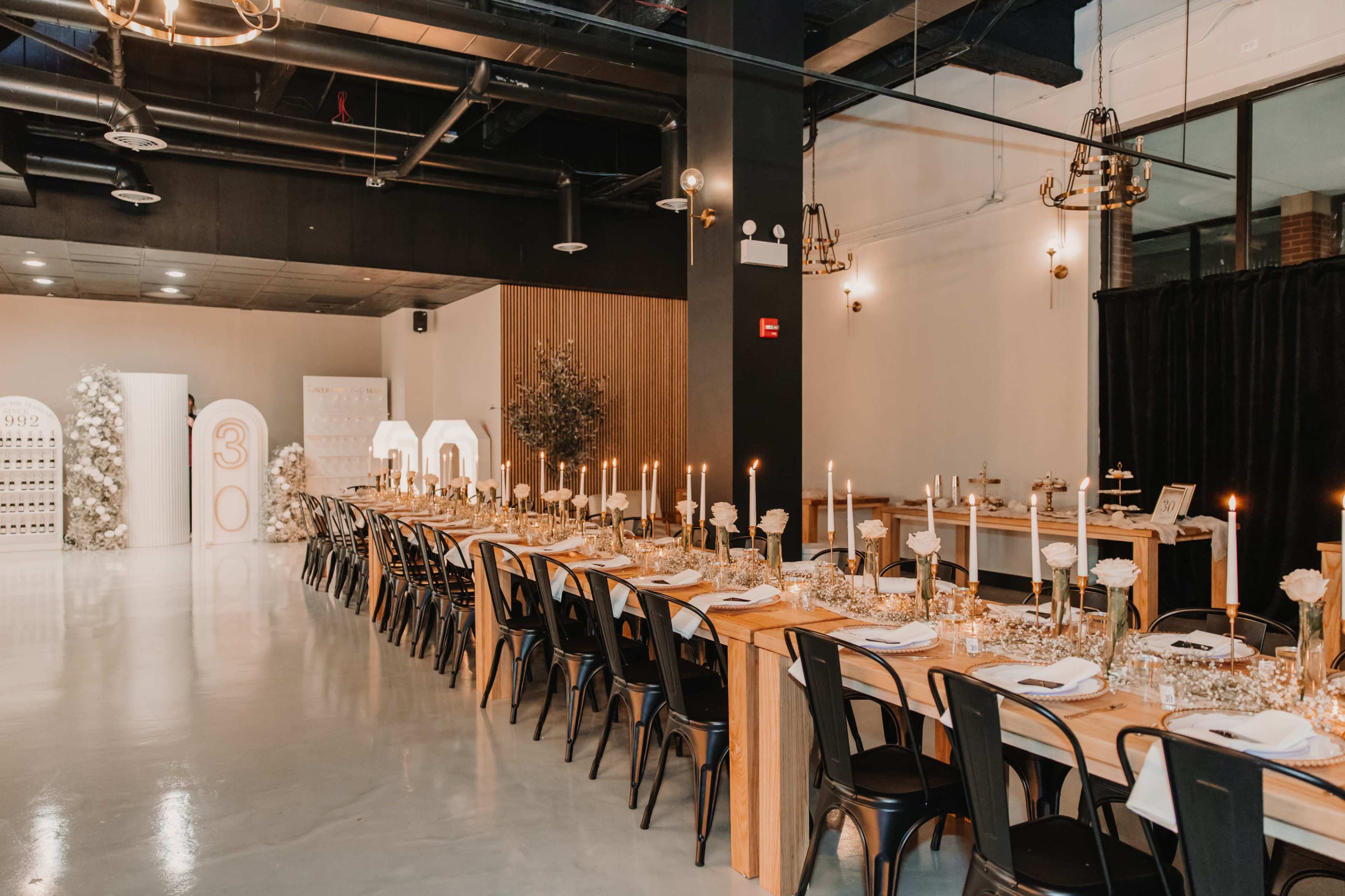 A long wooden dining table is set with white plates, candles, and floral centerpieces in a modern event space.