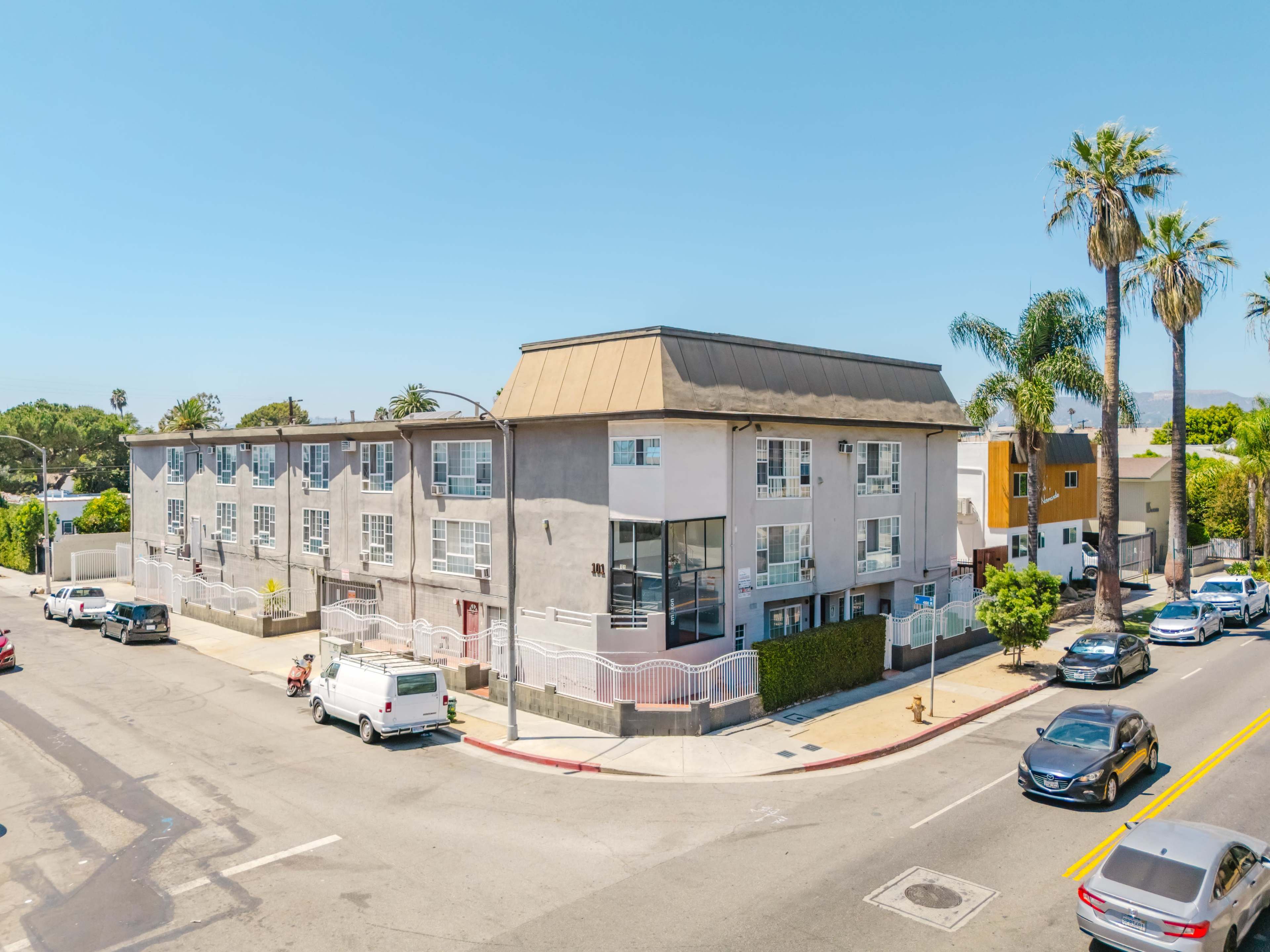 The image shows a three-story apartment building at the corner of a street, surrounded by palm trees and parked cars.