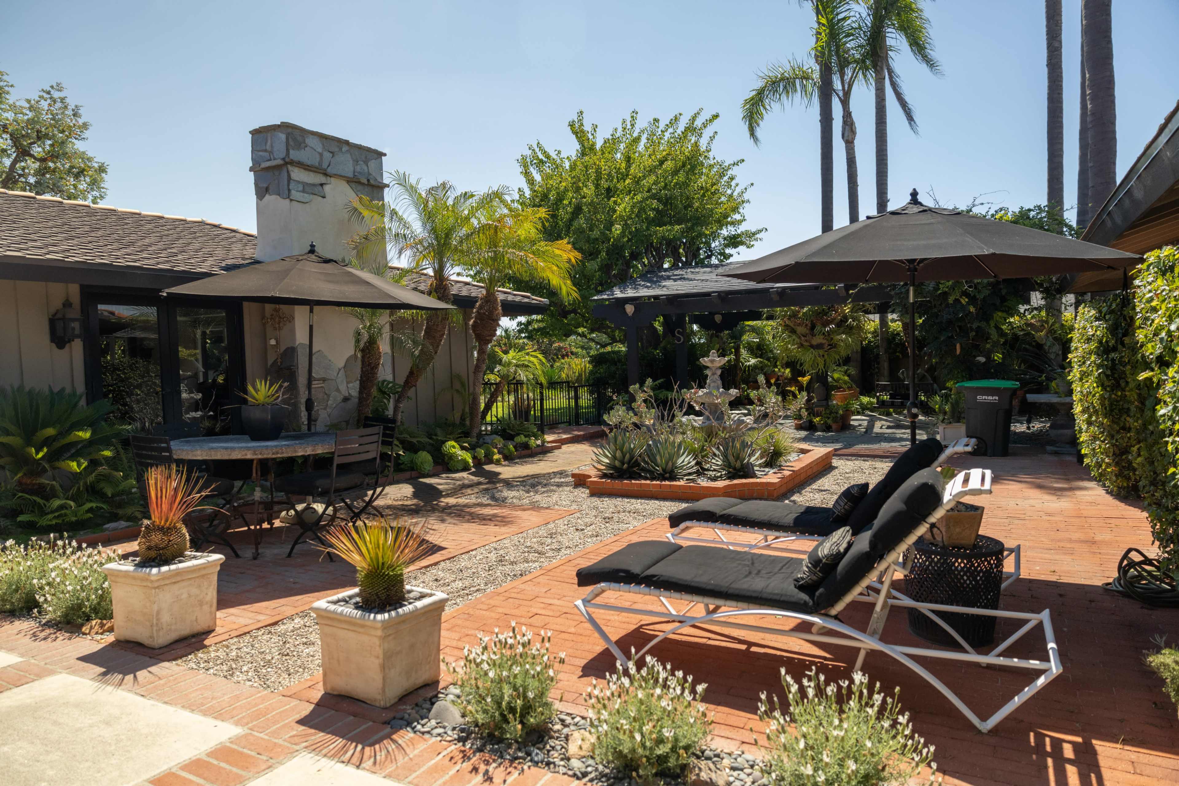 A landscaped backyard featuring lounge chairs, a dining table, and a decorative fountain surrounded by plants and palm trees.