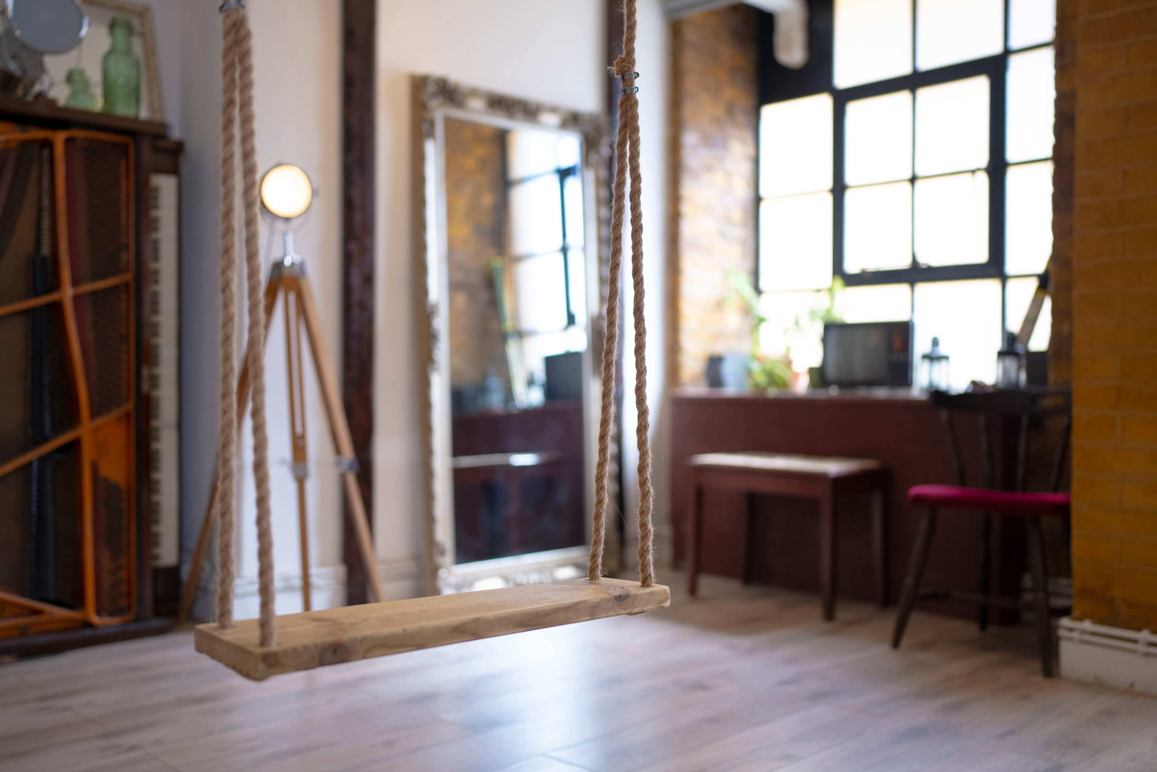 A wooden swing hanging from the ceiling in a room with large windows, a mirror, and various furniture pieces.