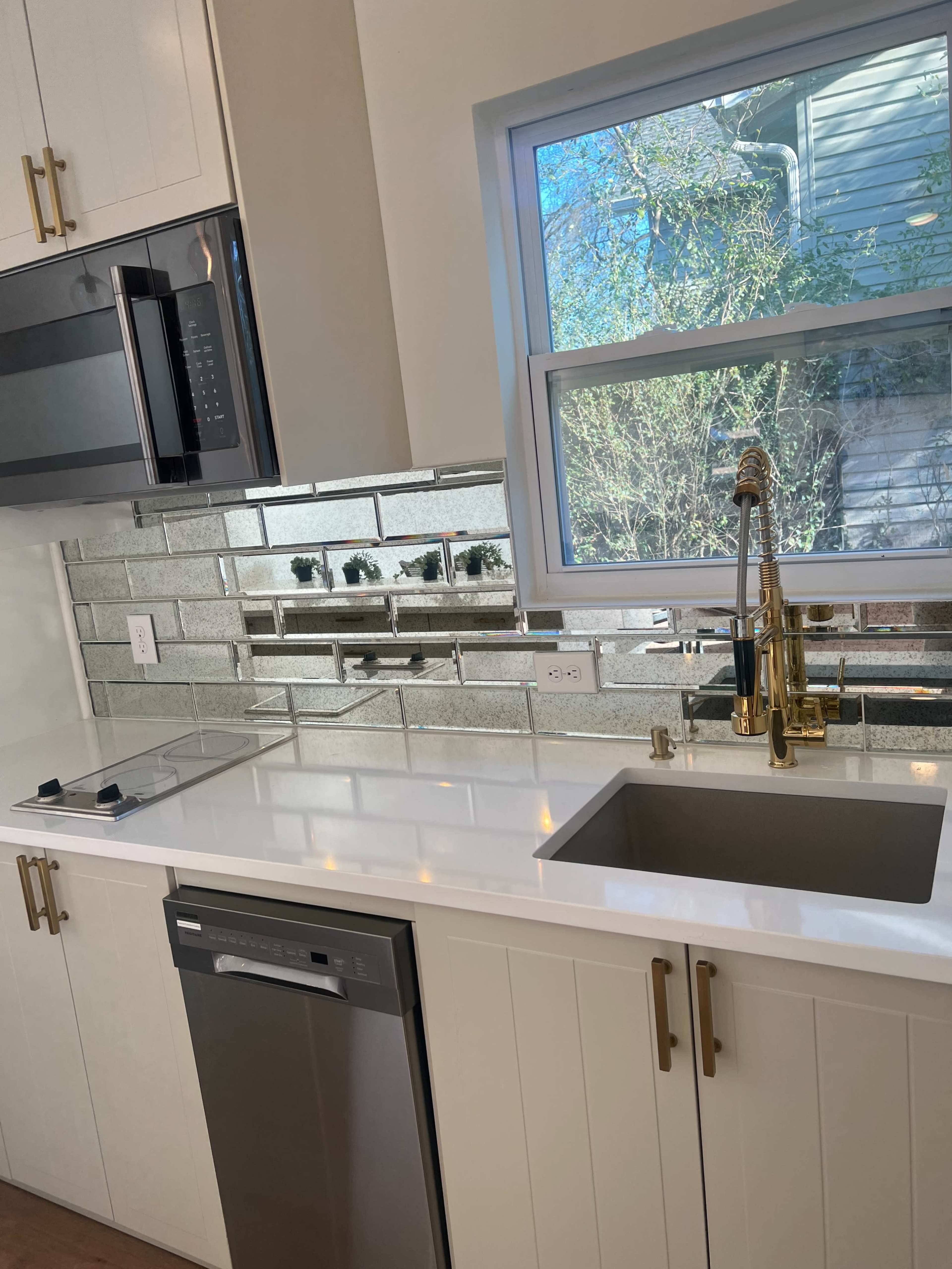 The image shows a modern kitchen with white cabinetry, a stainless steel dishwasher, a sleek sink, and a mirror-tiled backsplash.