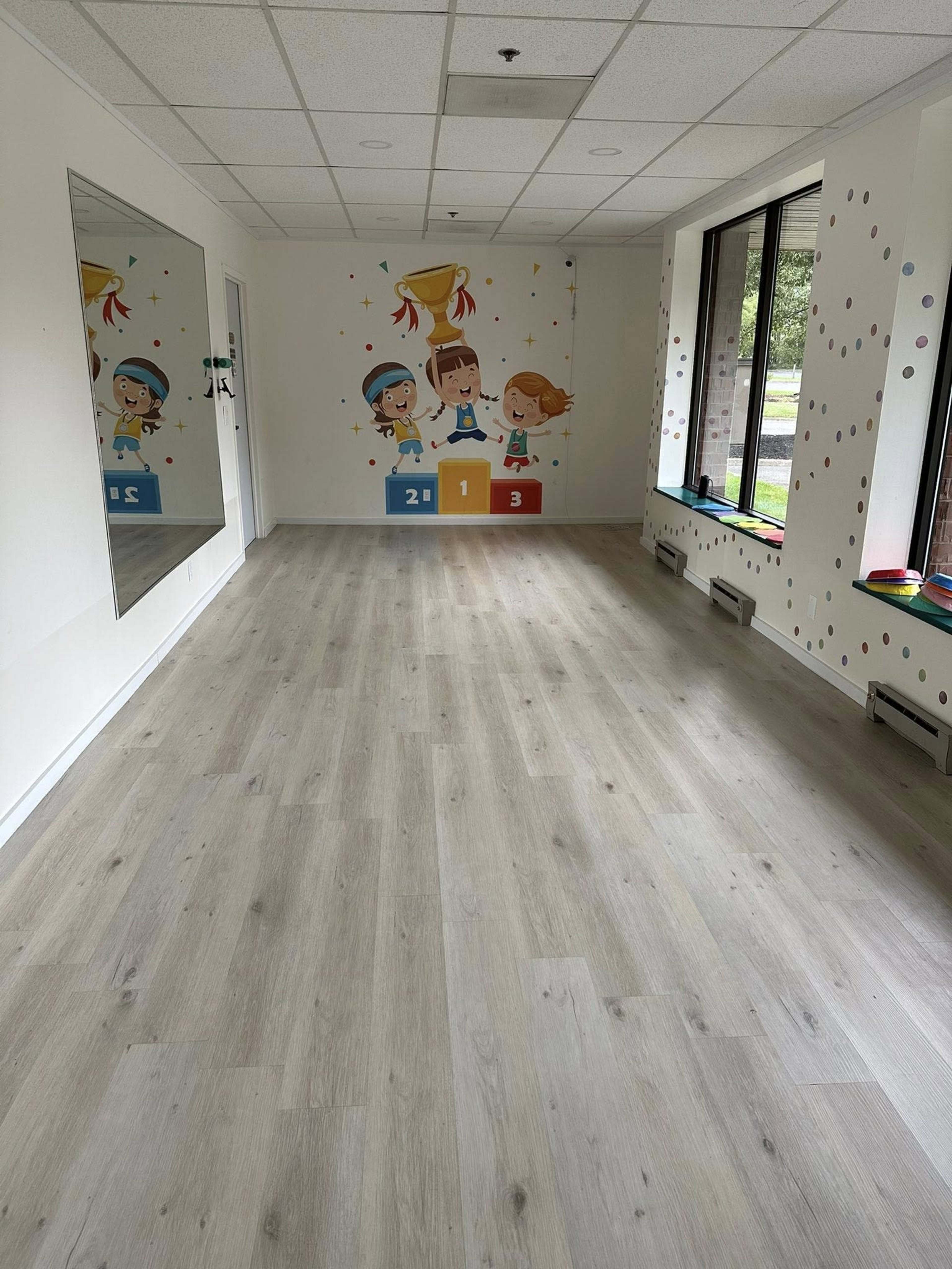 The image shows a bright, empty classroom with wooden flooring, decorated with colorful cartoon characters and numbers on the walls, and featuring large windows.