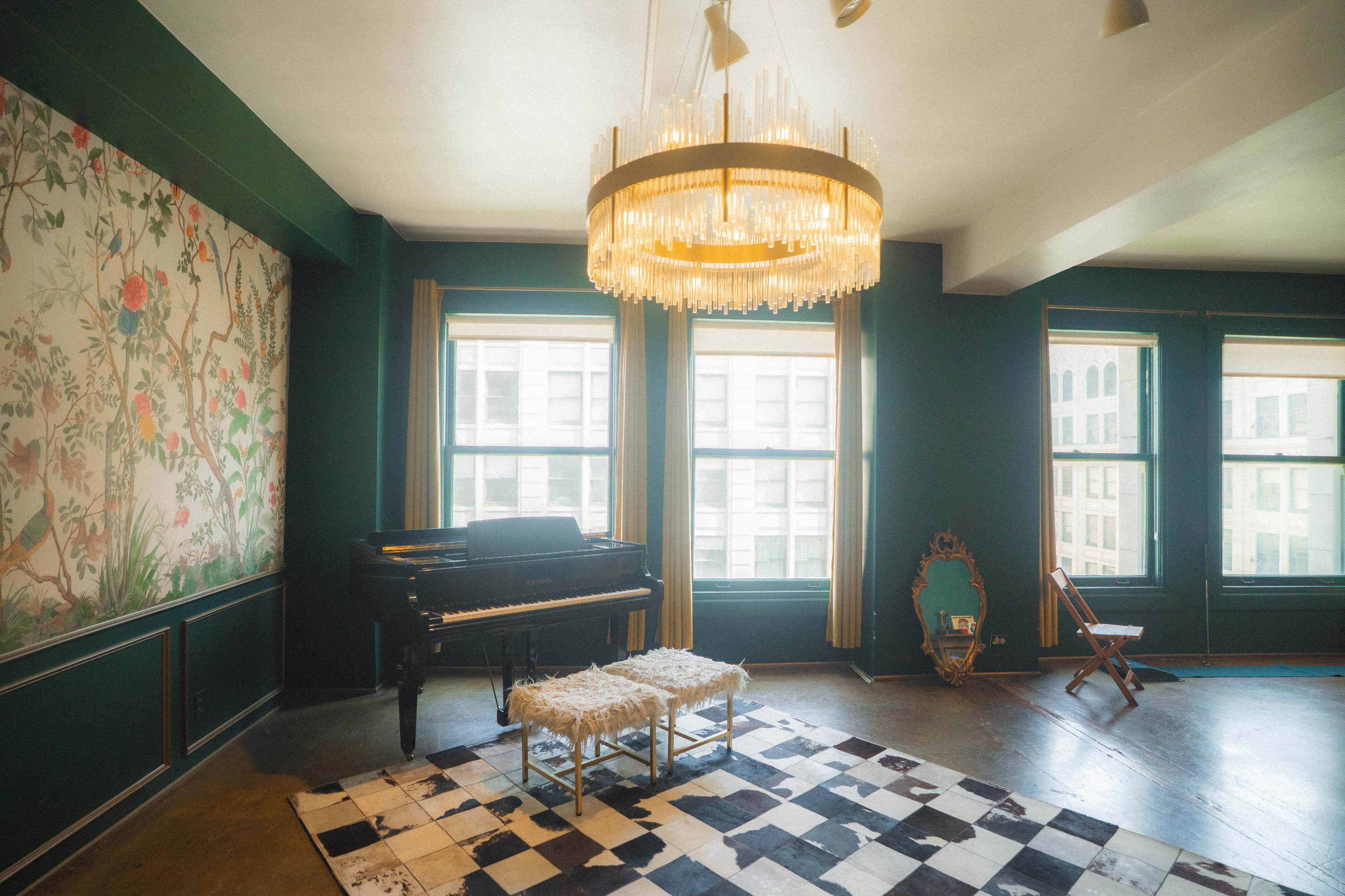 A spacious room features a black piano, two white stools, a large chandelier, and decorative wallpaper, with natural light streaming through the windows.