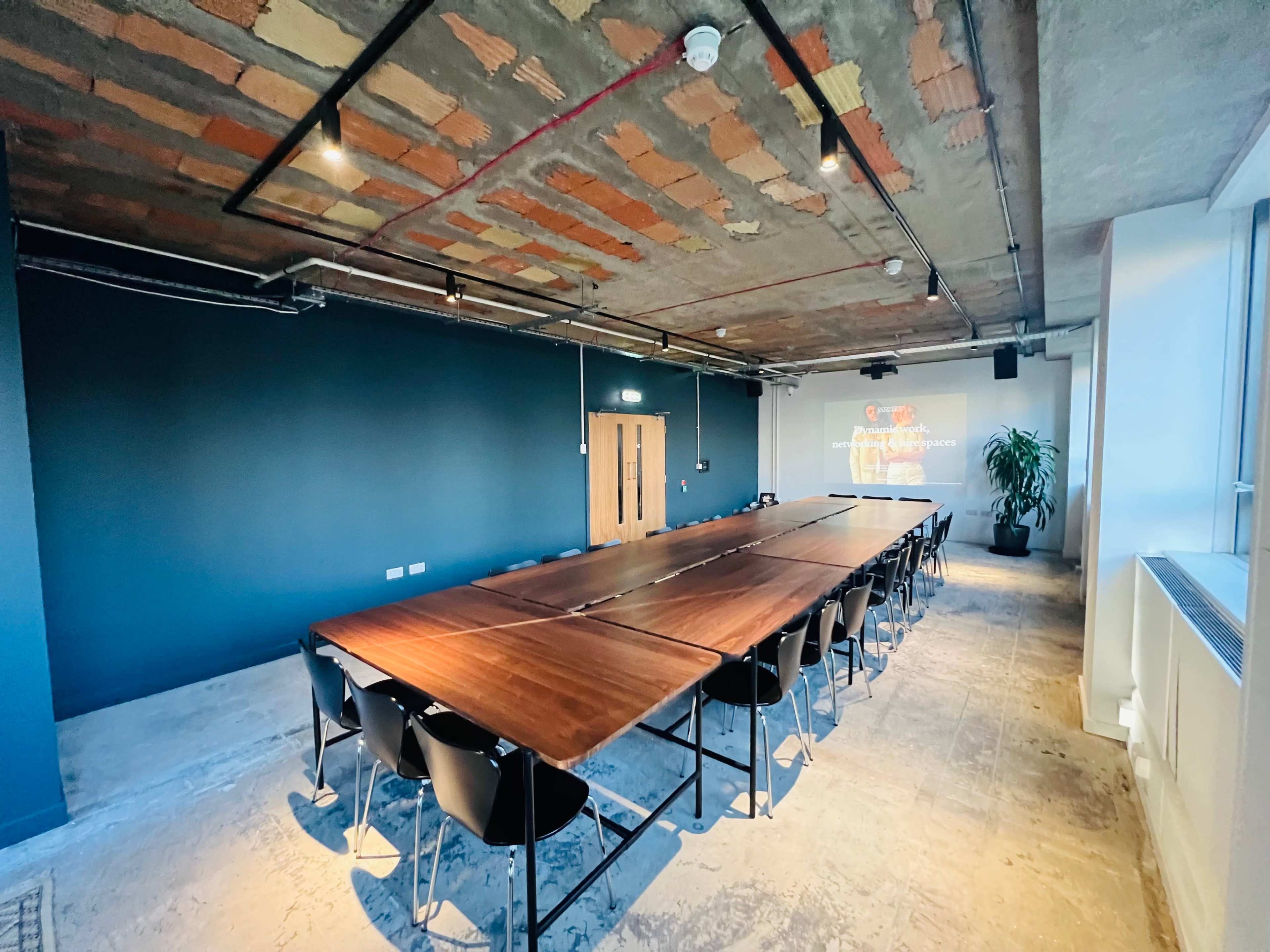 The image shows a spacious conference room featuring a long wooden table surrounded by black chairs, with a large wall projection on one side and exposed ceiling elements.