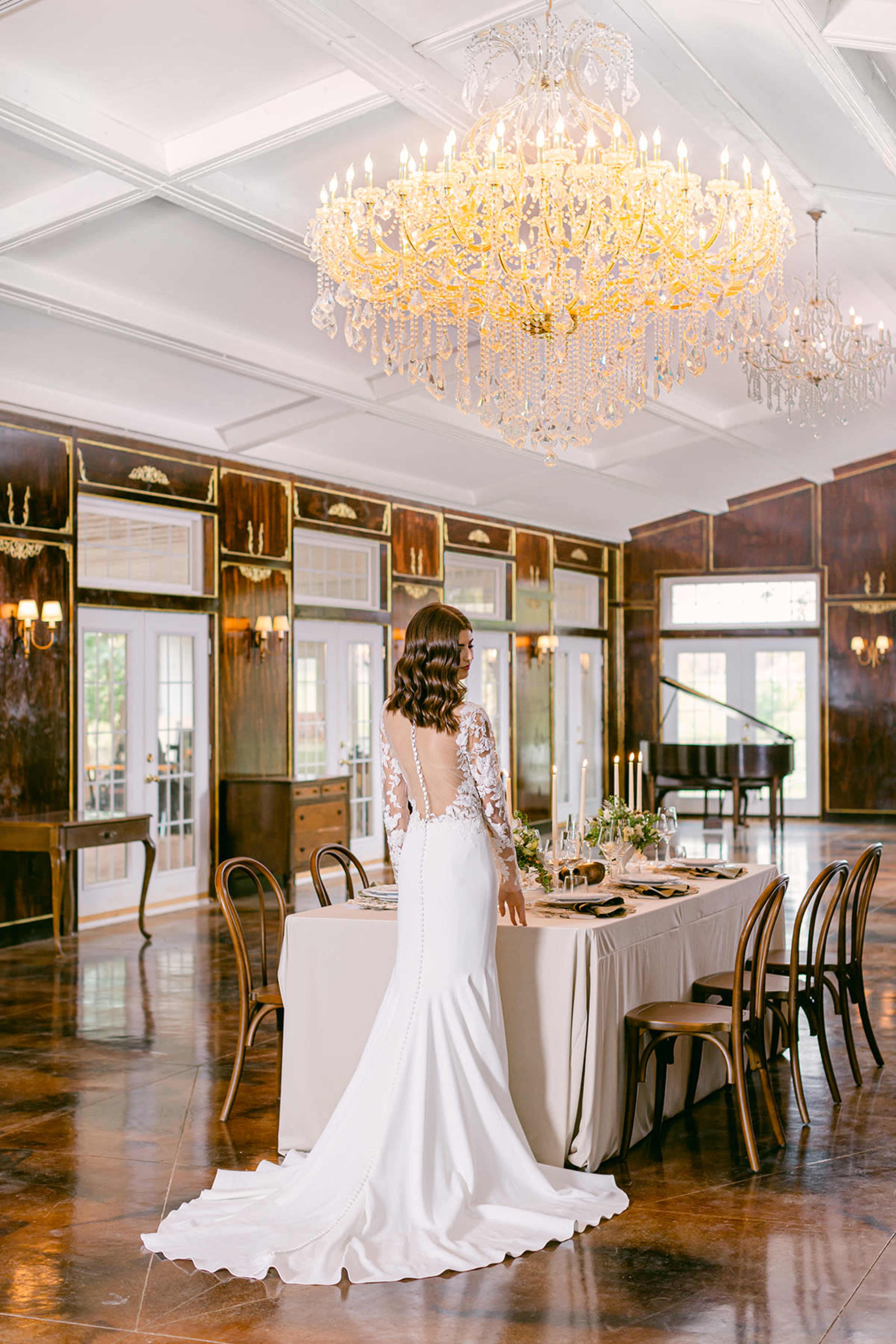 A woman stands in a beautifully arranged dining room with a grand chandelier and an elegant table setup.