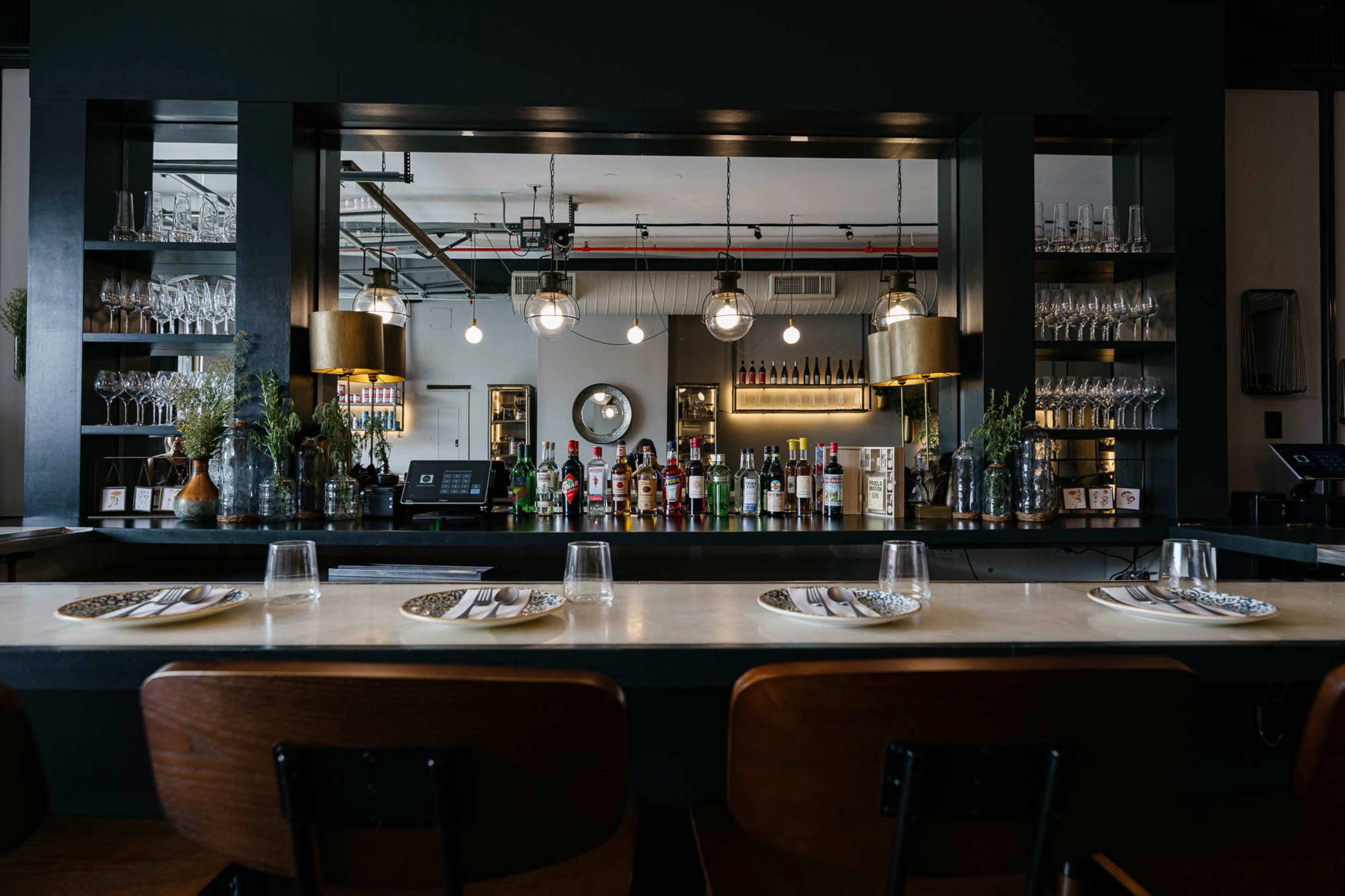 The image shows a modern bar area with a countertop, seating, and a variety of bottles and glassware displayed on shelves in a dimly lit environment.