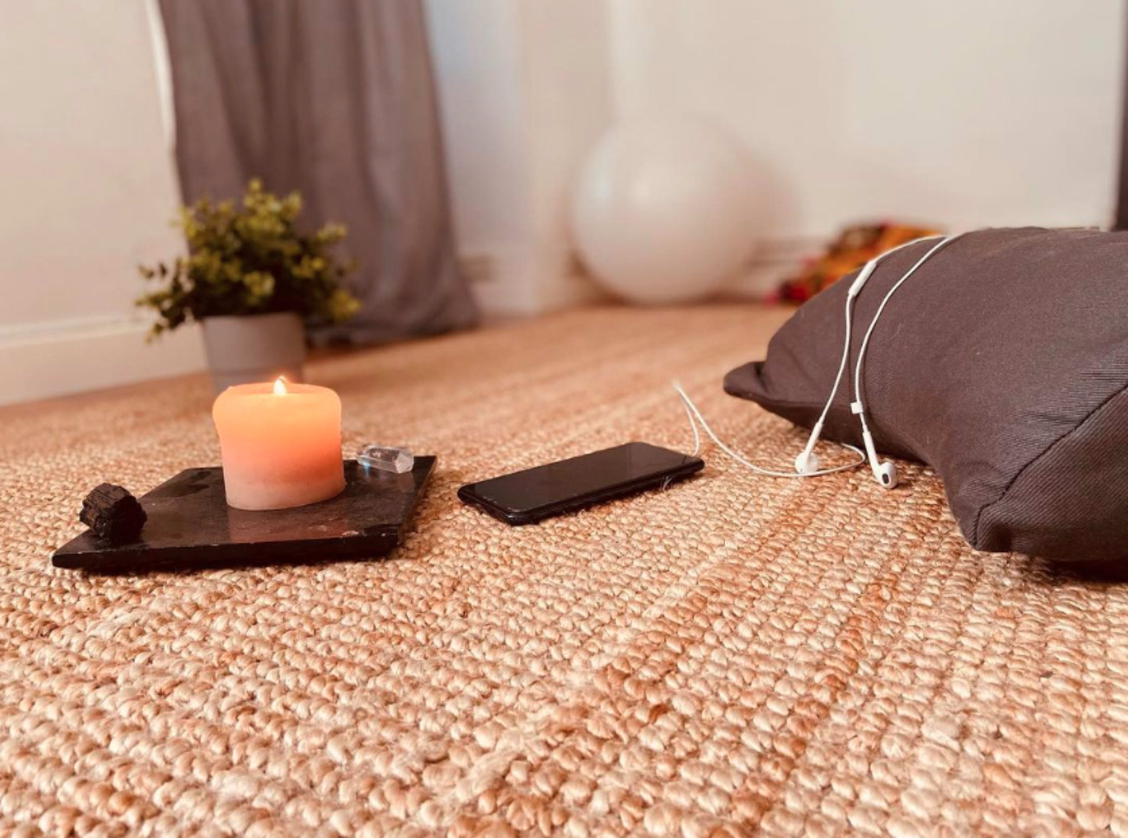 A lit candle sits on a black plate next to a smartphone with earphones on a textured woven floor beside a cushion and a potted plant.