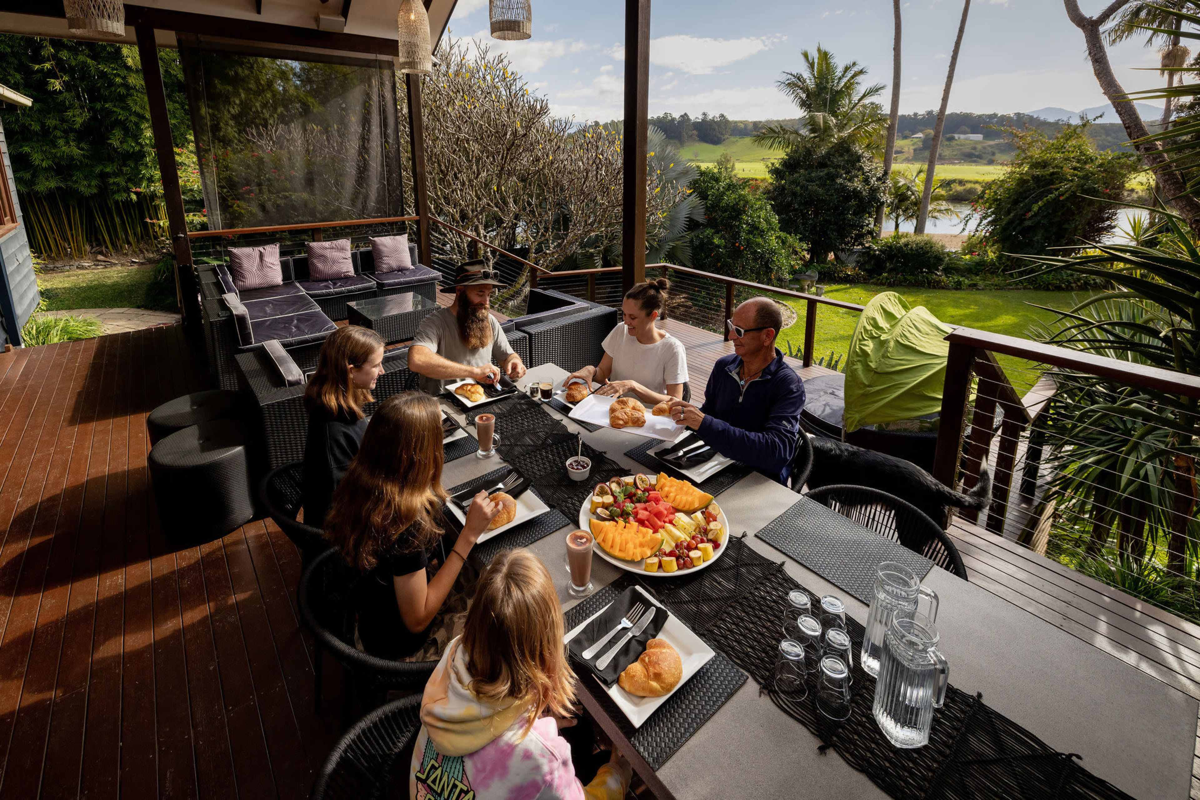 A group of six people enjoys a meal on a patio surrounded by greenery and a view of a lake.