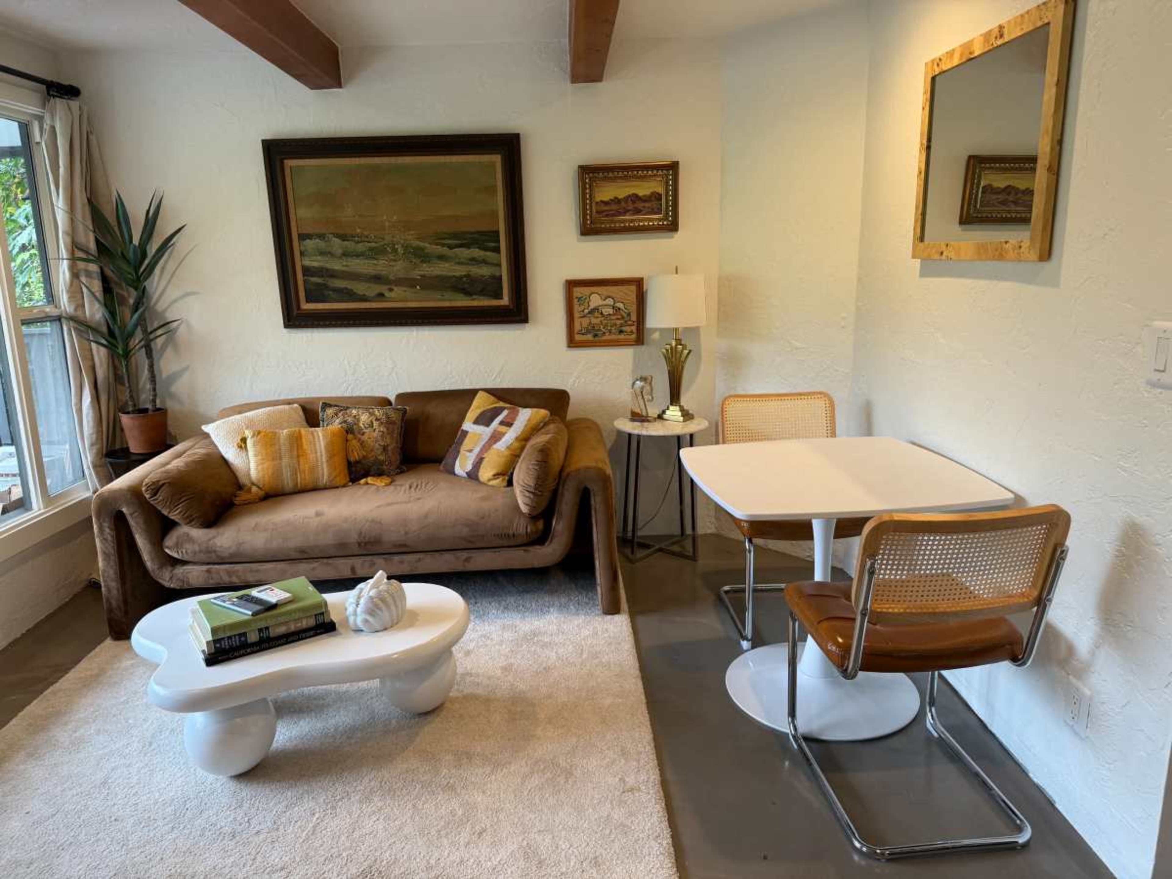 The image shows a cozy living space featuring a brown sofa with decorative pillows, a small white coffee table, a dining table with two chairs, and a large window allowing natural light to enter.