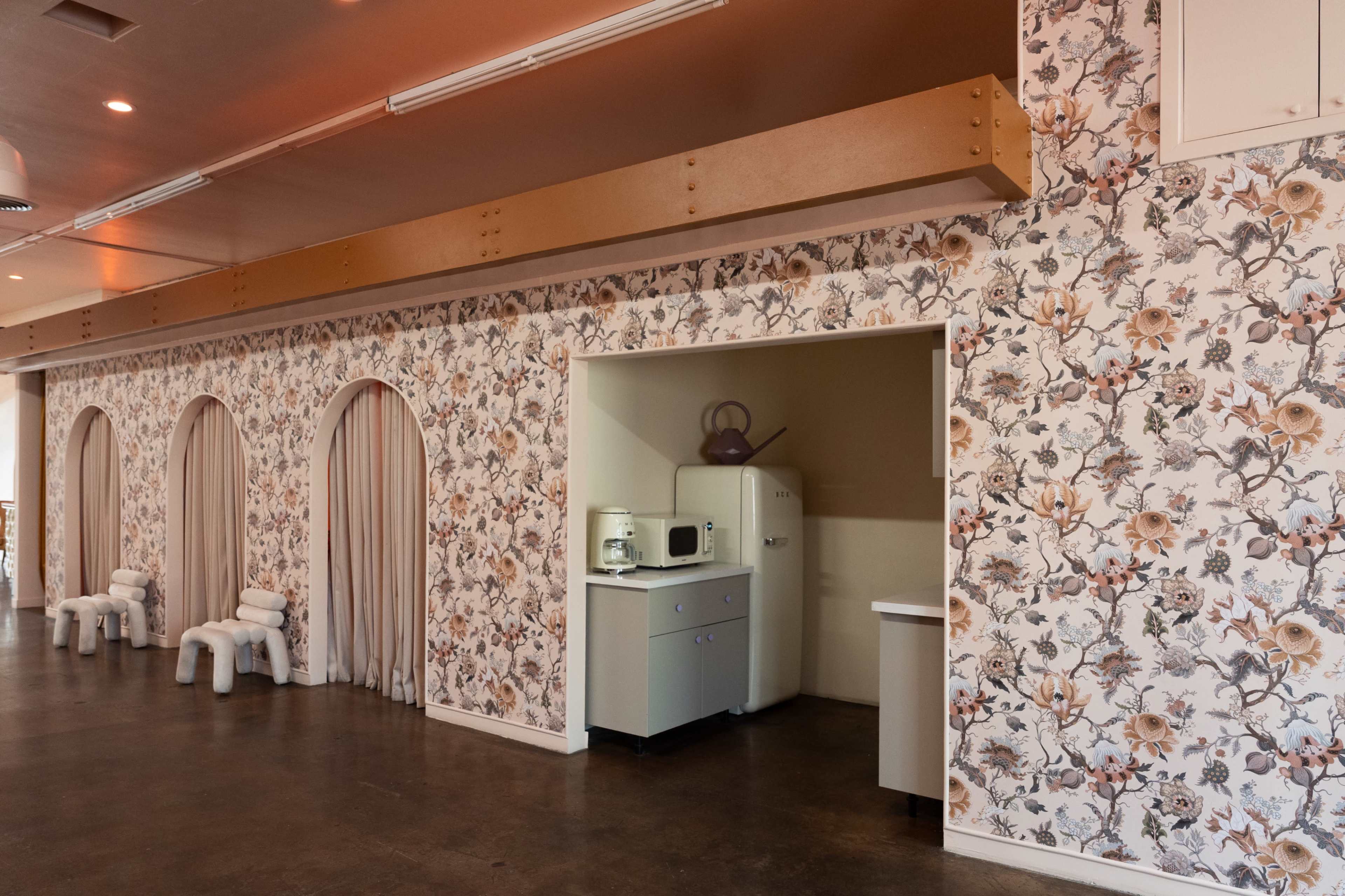 The image shows an interior space with floral wallpaper, archways draped in curtains, and a kitchenette area featuring a small refrigerator.