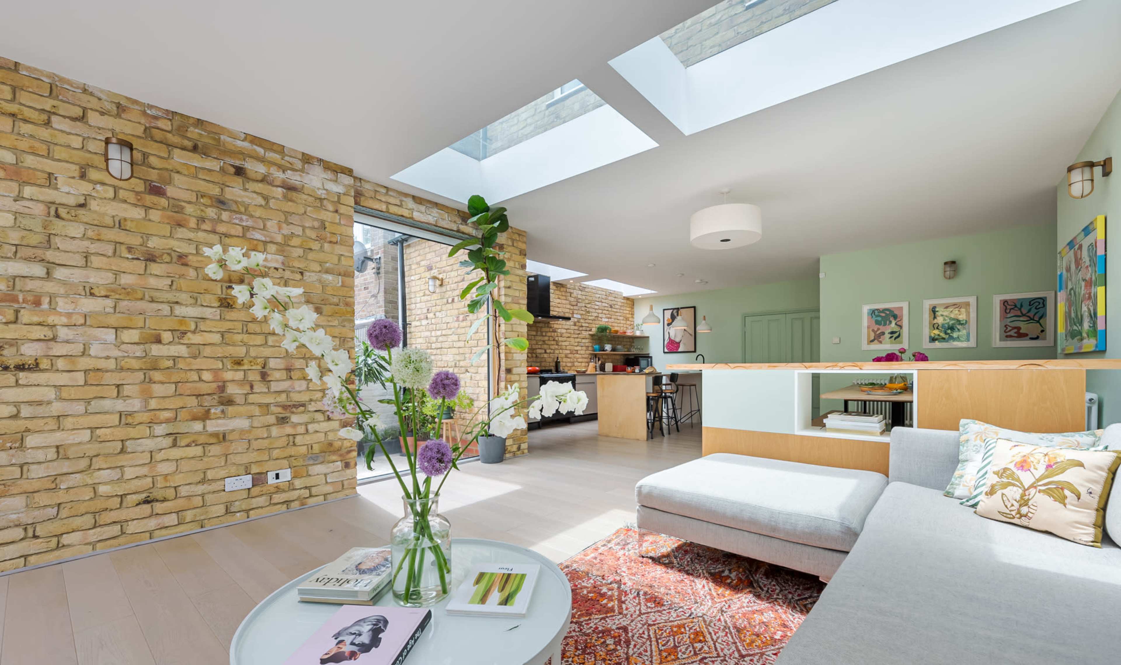 The image features a bright living room with brick walls, large skylights, and a sectional sofa, complemented by indoor plants and a modern coffee table.
