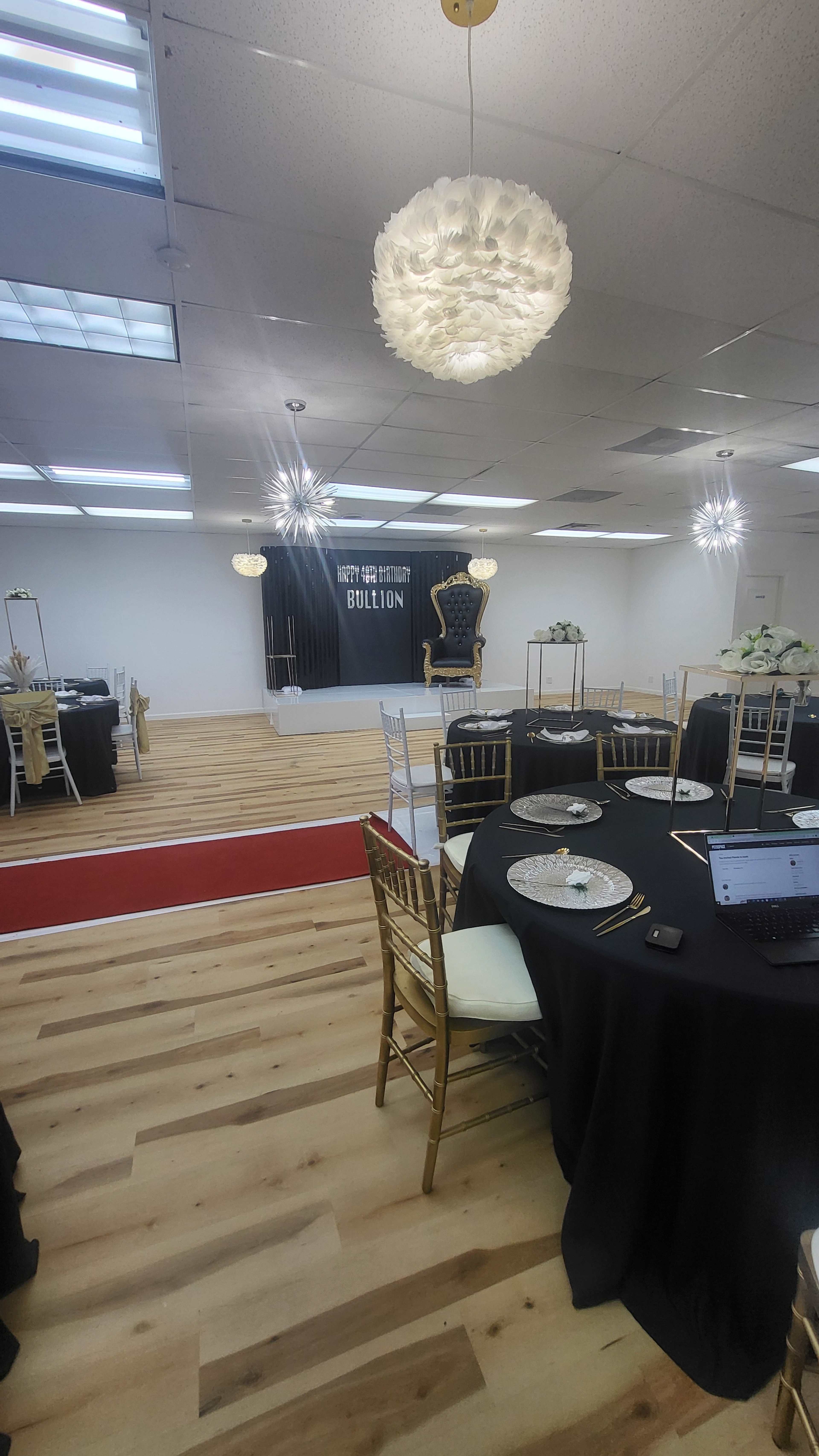 A spacious event hall with round tables set for dining, a red carpet leading to a decorated backdrop, and elegant lighting fixtures on the ceiling.
