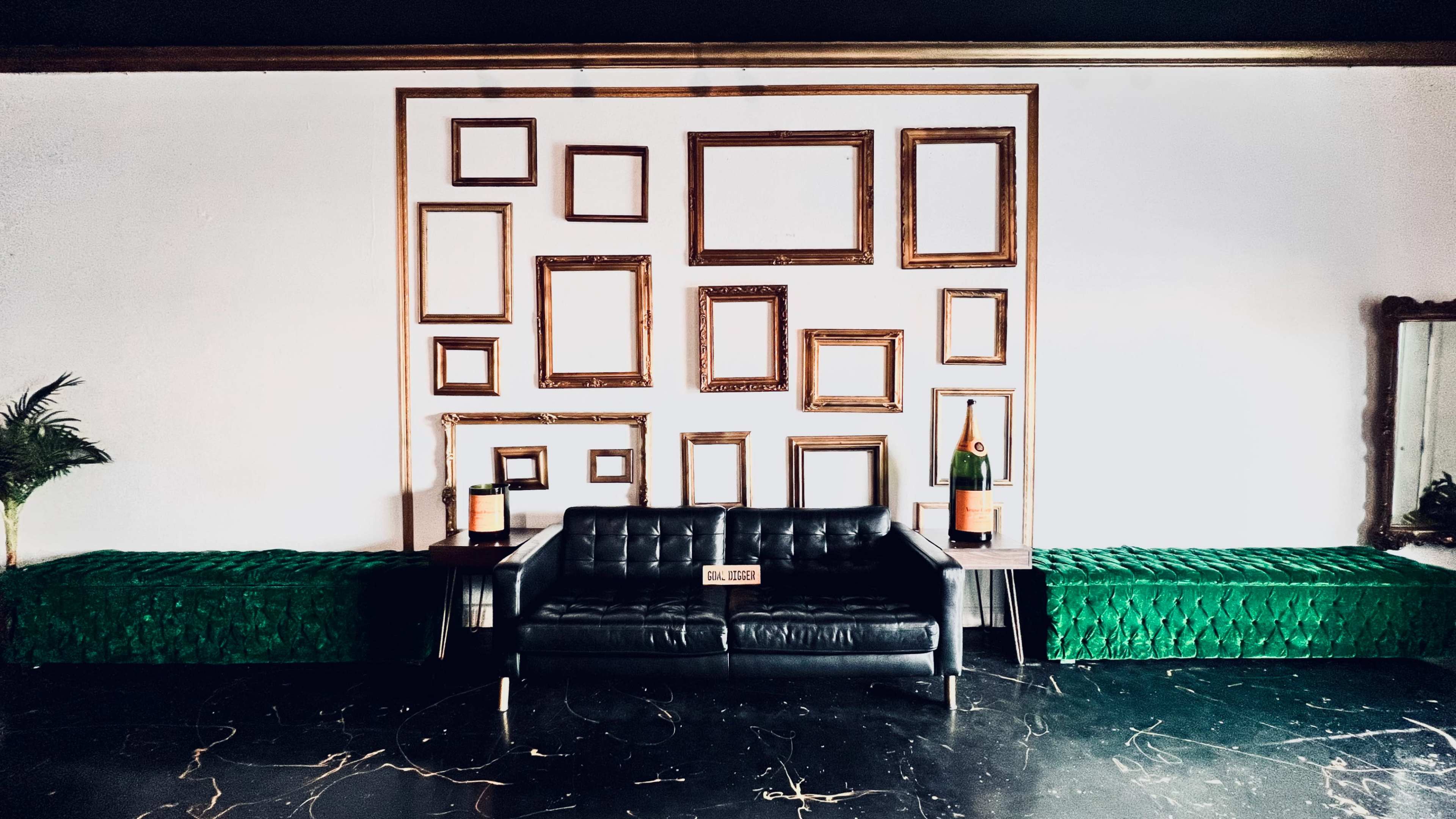 A black leather sofa is positioned in front of a wall decorated with various empty, ornate gold-framed picture frames, accompanied by two green benches.