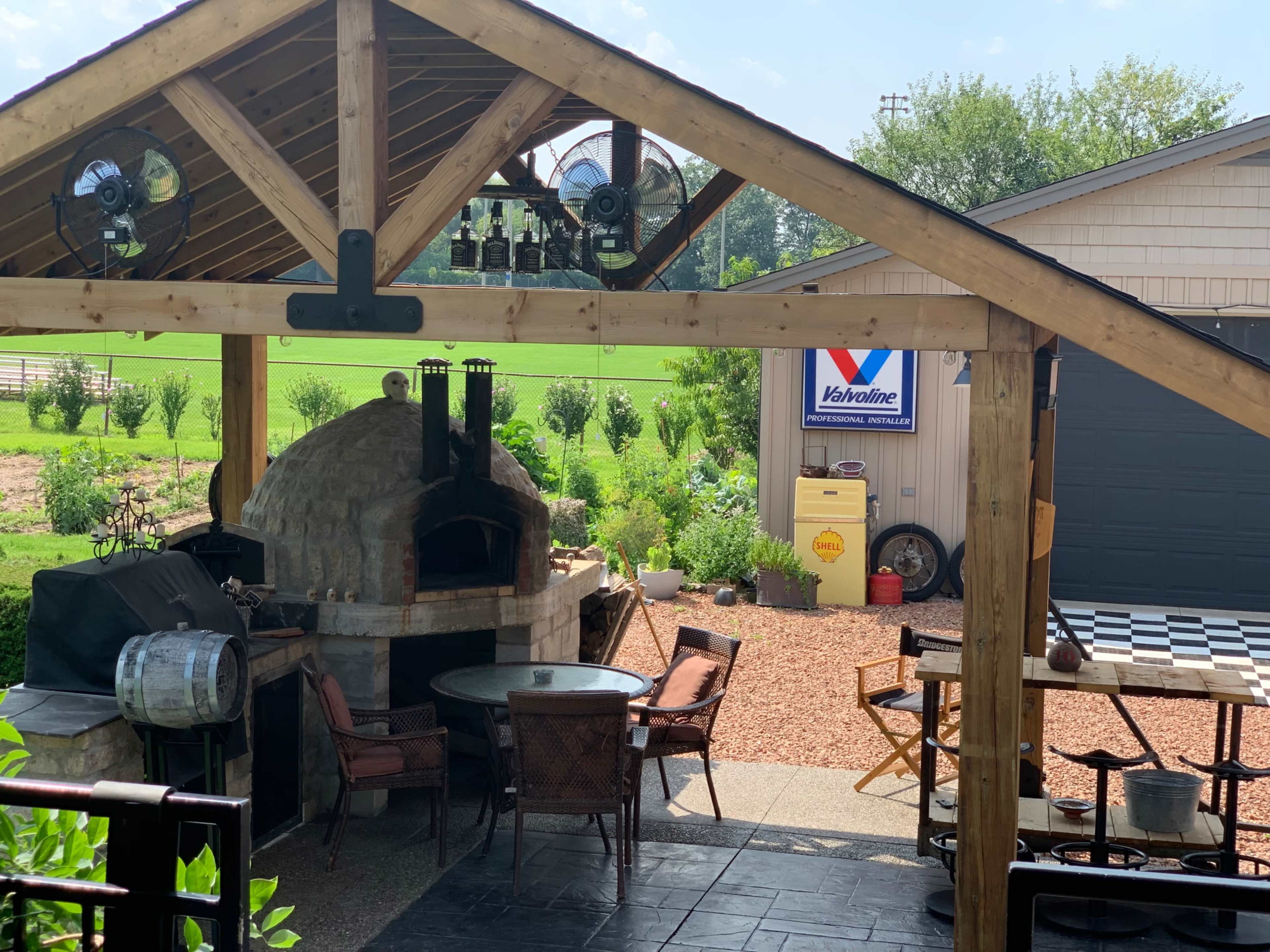 An outdoor patio featuring a stone pizza oven, a grill, and a seating area under a wooden pergola.
