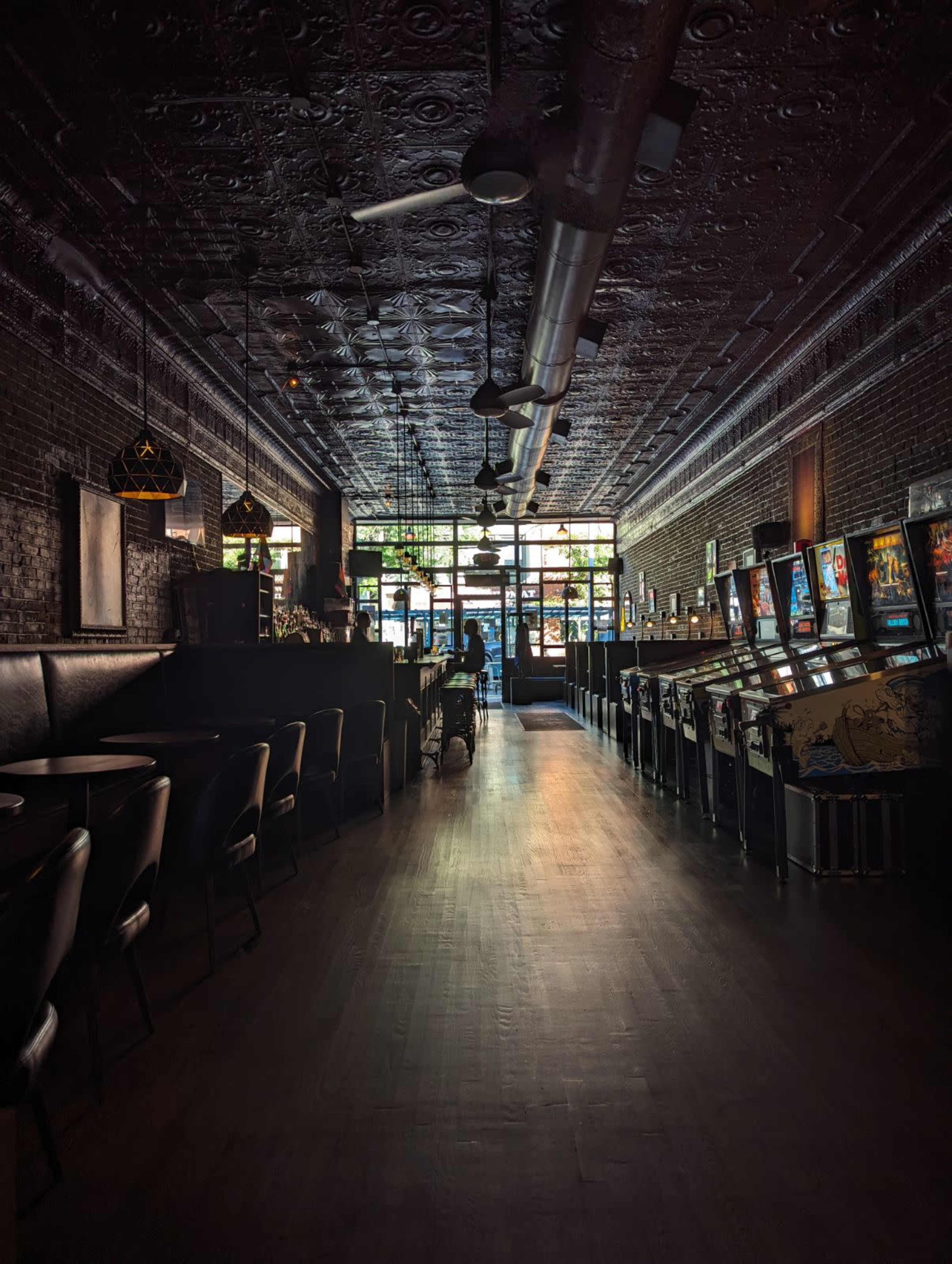 The image depicts a dimly lit bar or arcade with rows of vintage arcade machines lining the walls and a large window allowing natural light to enter.