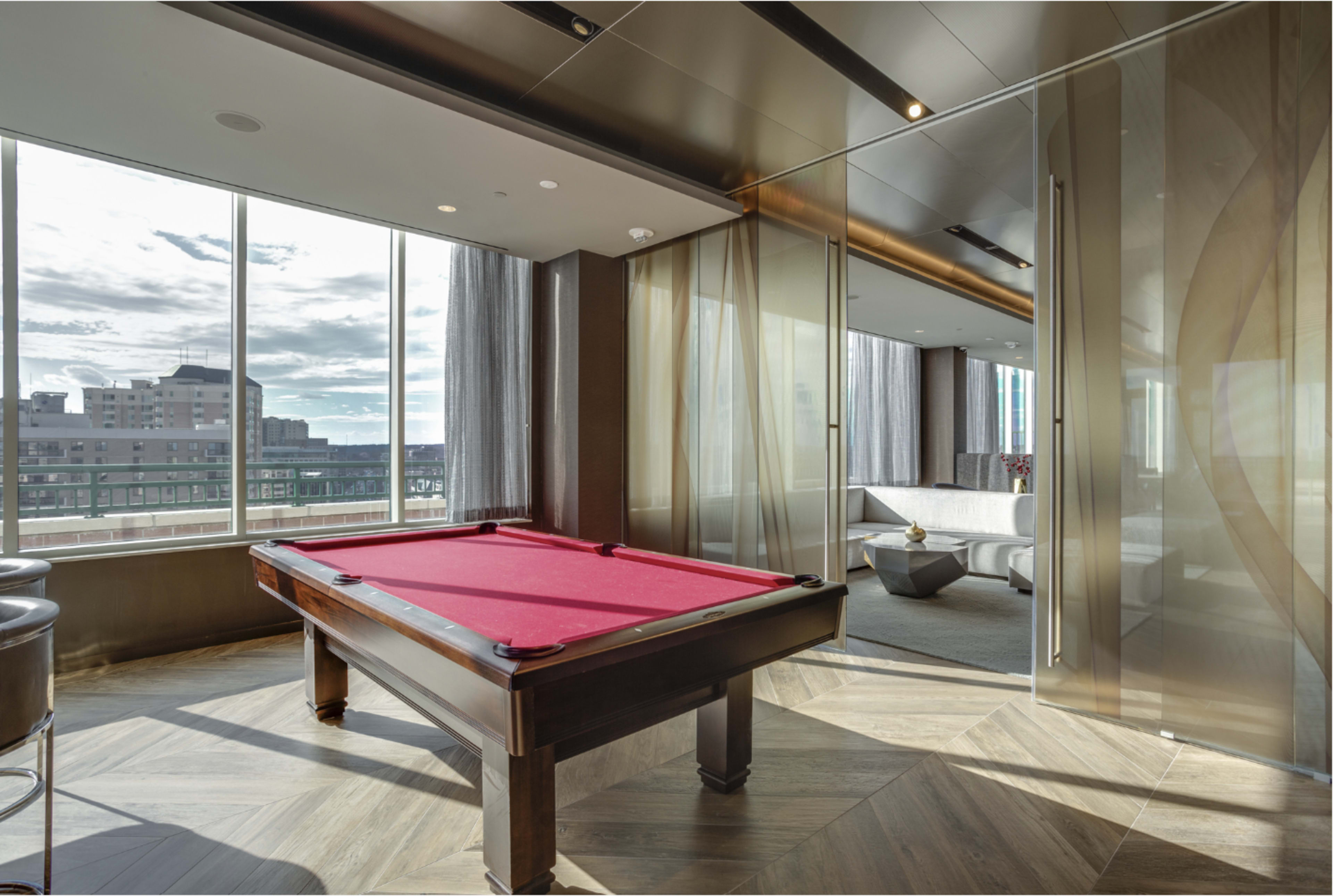 A pool table with a red felt surface is positioned near large windows offering a view of an urban skyline.