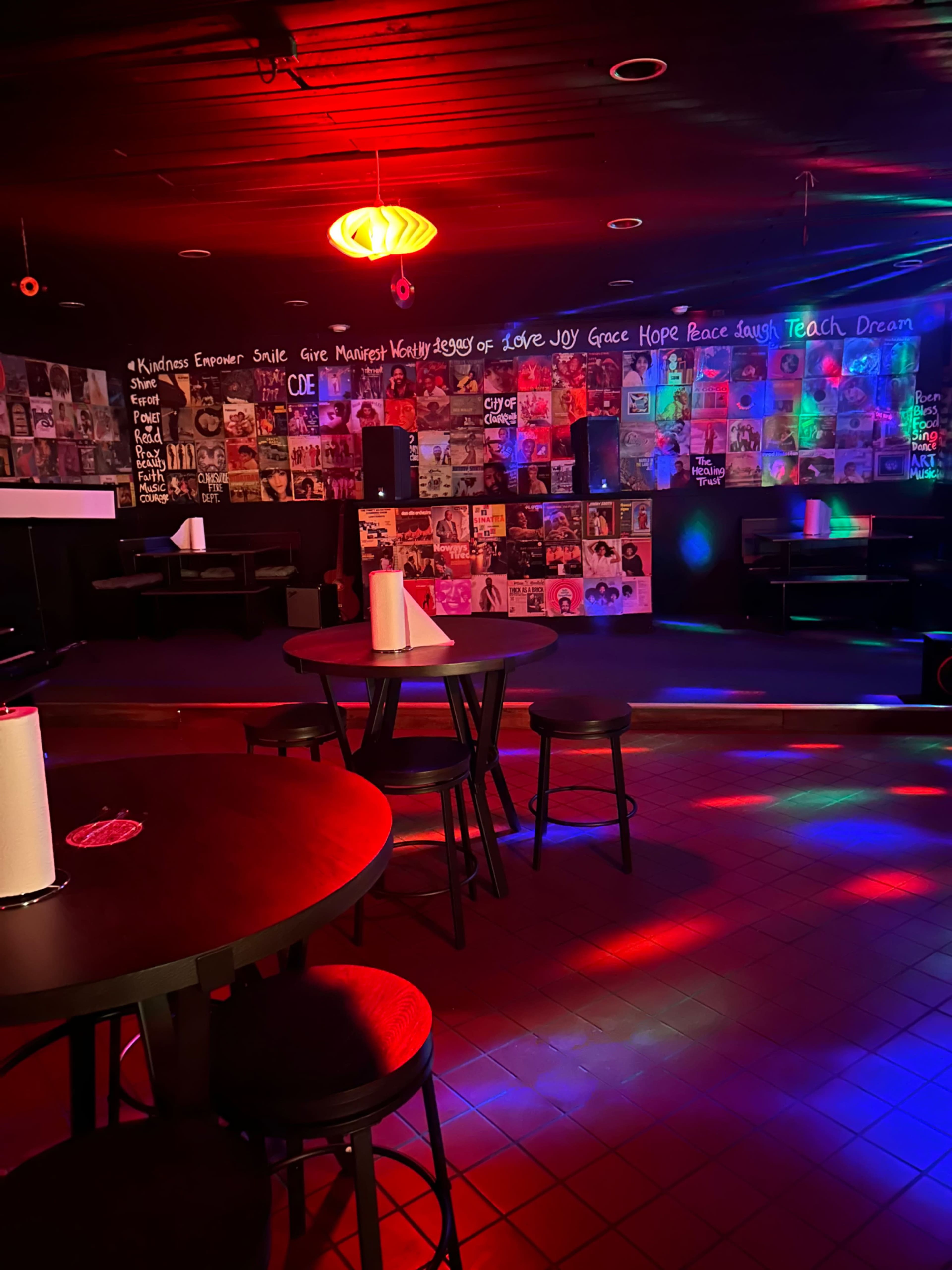 An underground bar with dim lighting, patterned walls covered in posters, and several tables with black chairs.