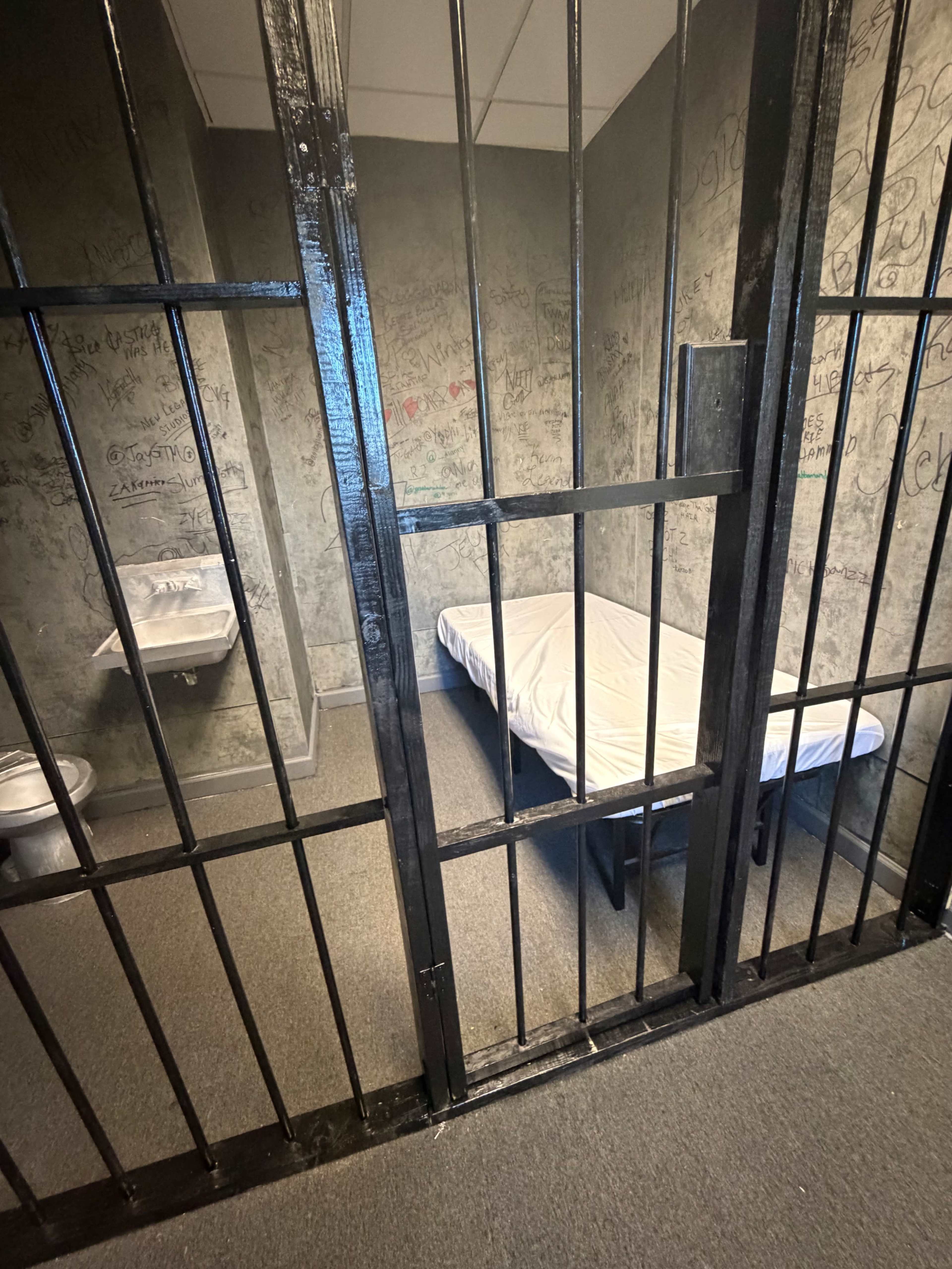 The image shows a small jail cell with a metal bed frame, a mattress, a toilet, and graffiti-covered walls.