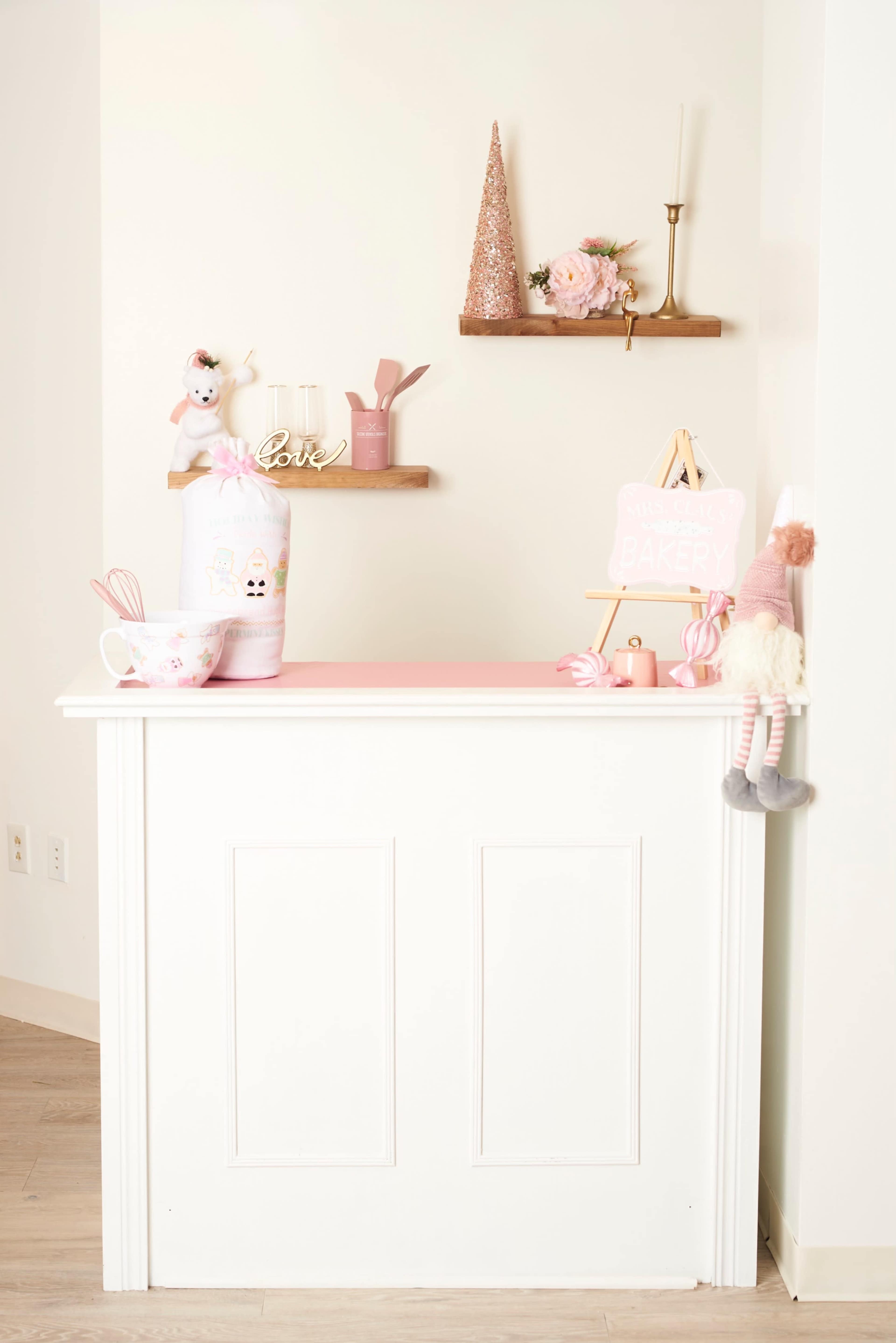 A small, decorated counter with a pink top, a decorative sign for a bakery, a mixing bowl, and various pink-themed items displayed on wooden shelves.