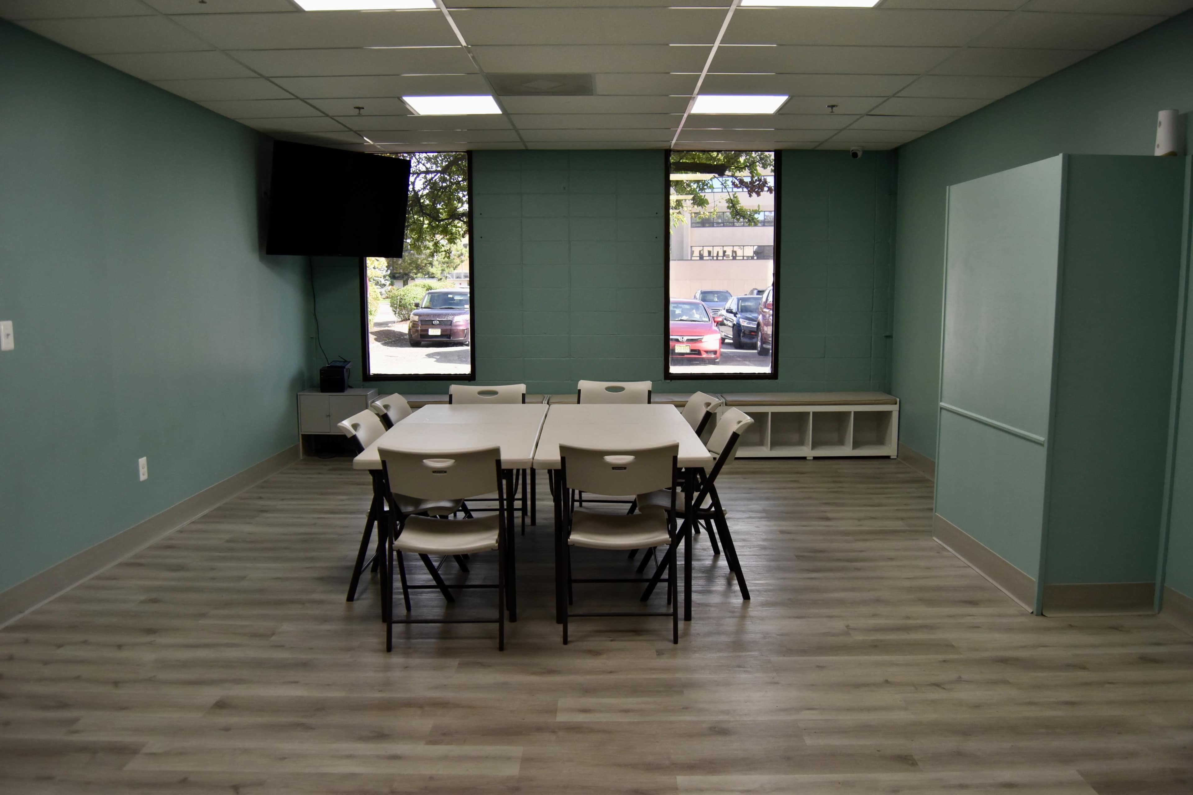 A conference room features a central table surrounded by chairs, with a television mounted on the wall and large windows providing outdoor views.