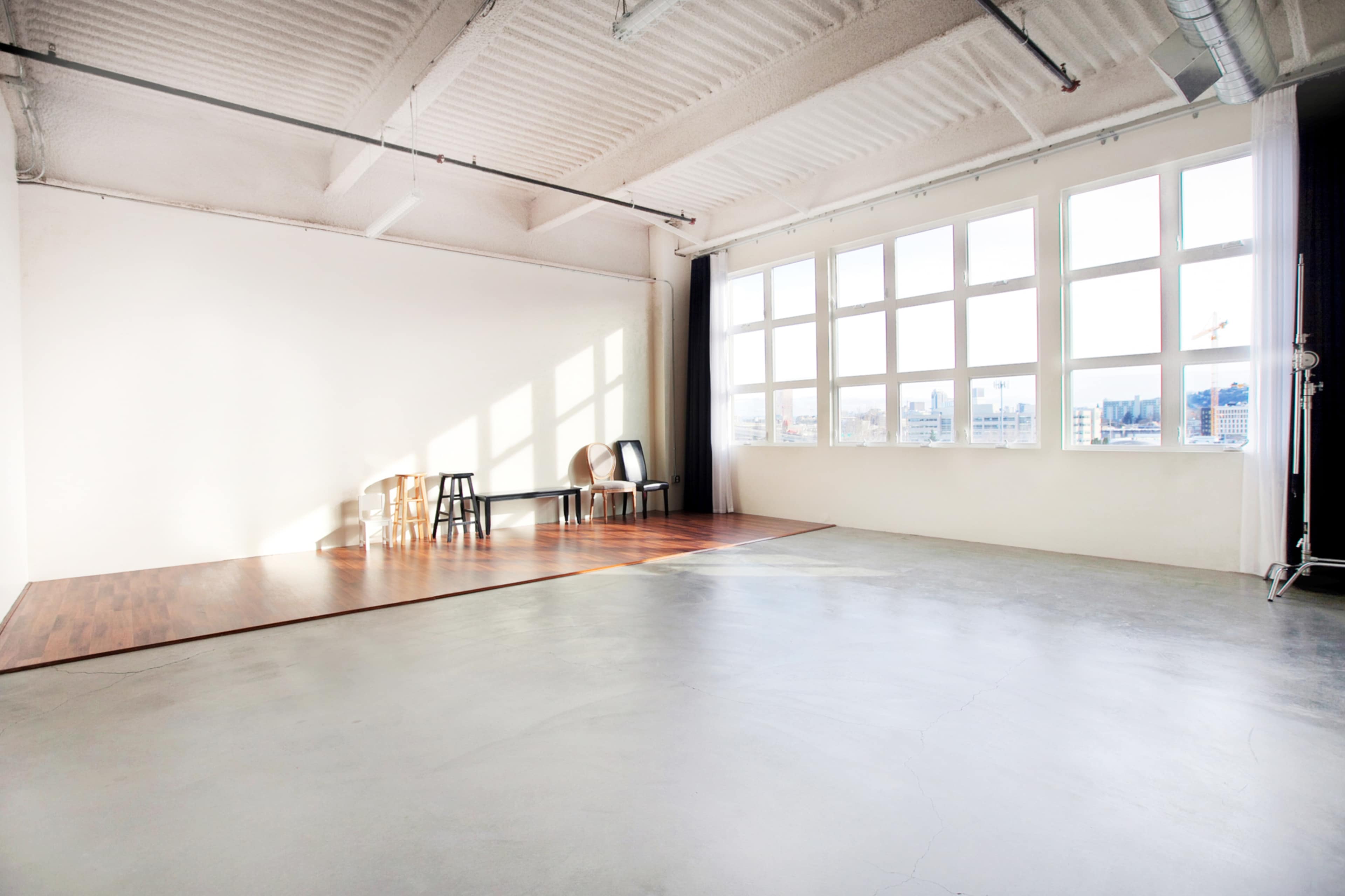 The image shows a spacious studio with large windows, wooden flooring in one section, and a few chairs positioned near the wall.