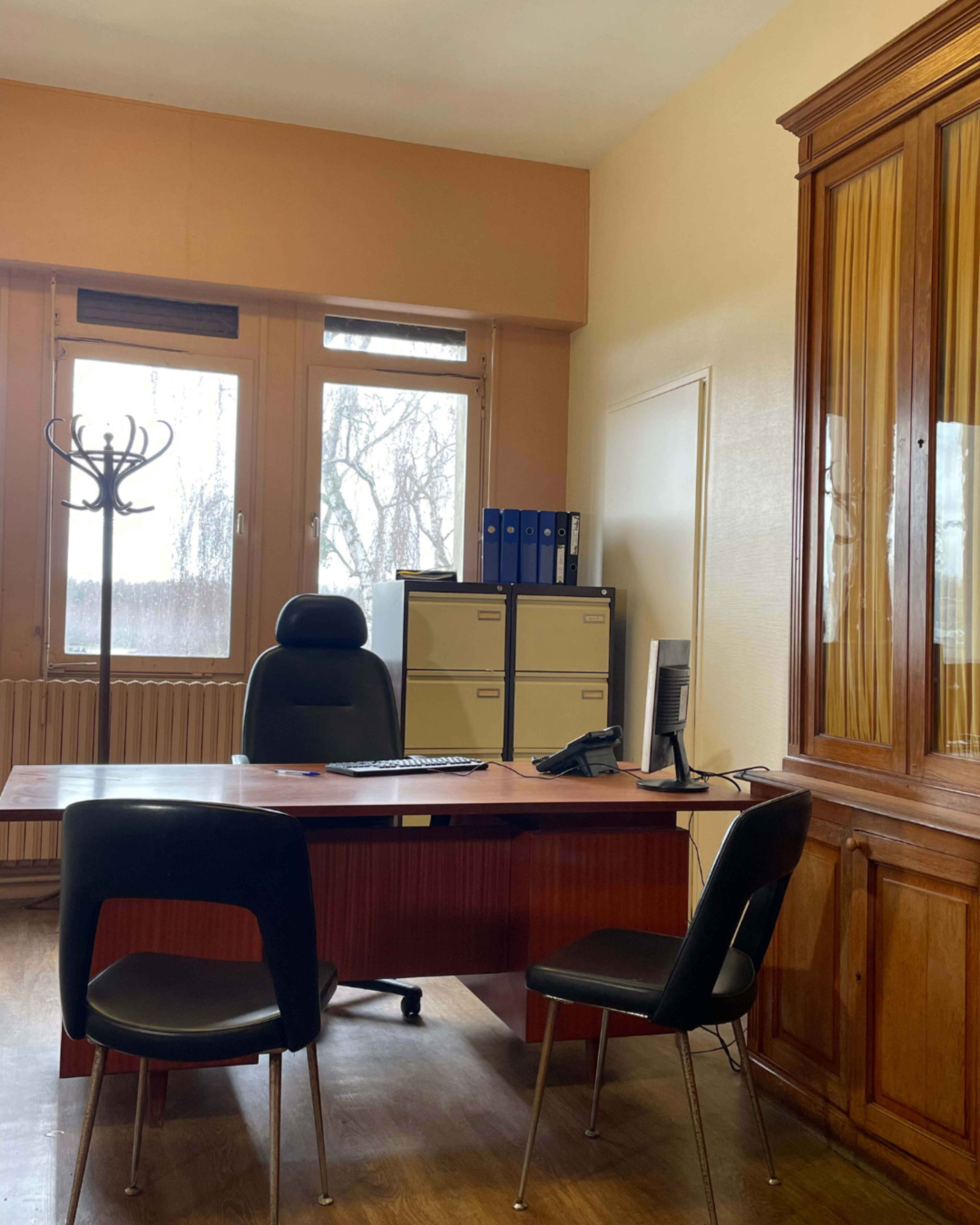 An office with a wooden desk, an ergonomic chair, two black chairs, a computer monitor, a filing cabinet, and a coat rack, illuminated by natural light from large windows.