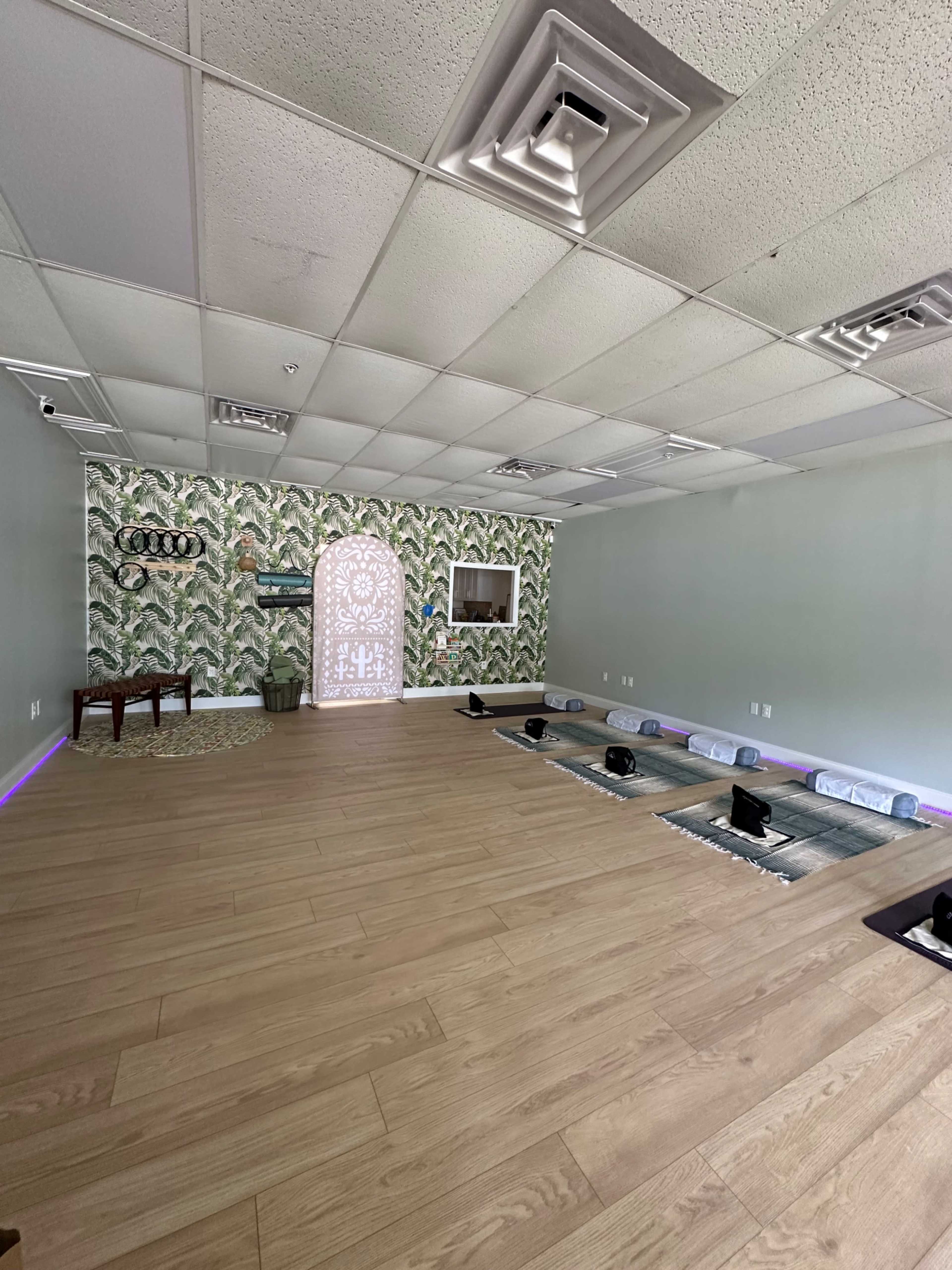 The image shows a spacious, minimalist room with hardwood flooring, adorned with a decorative wall featuring leafy patterns, and arranged with seating cushions facing a decorative archway.