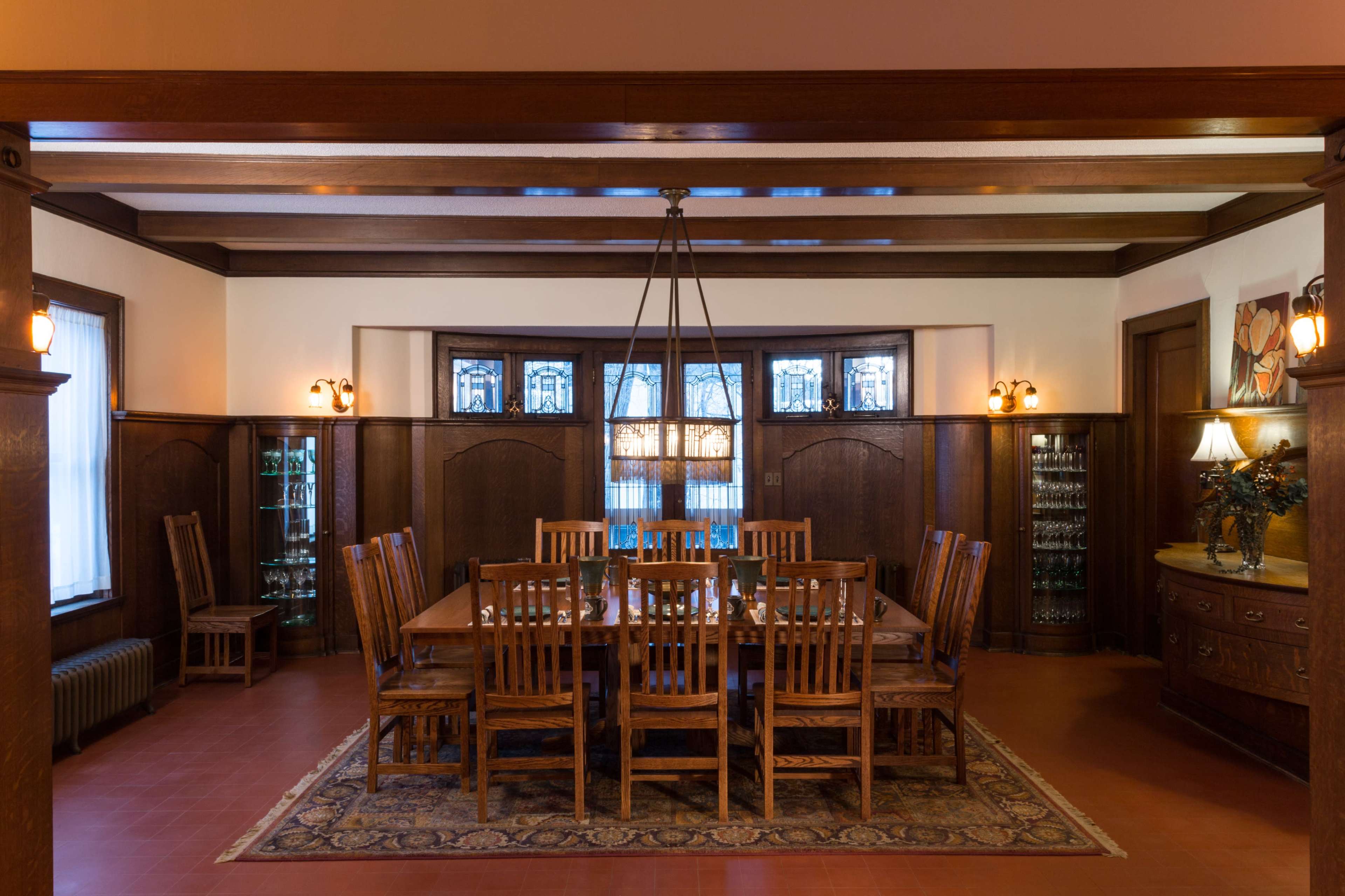 A spacious dining room with a large wooden table surrounded by chairs, illuminated by pendant lighting and featuring stained glass windows.