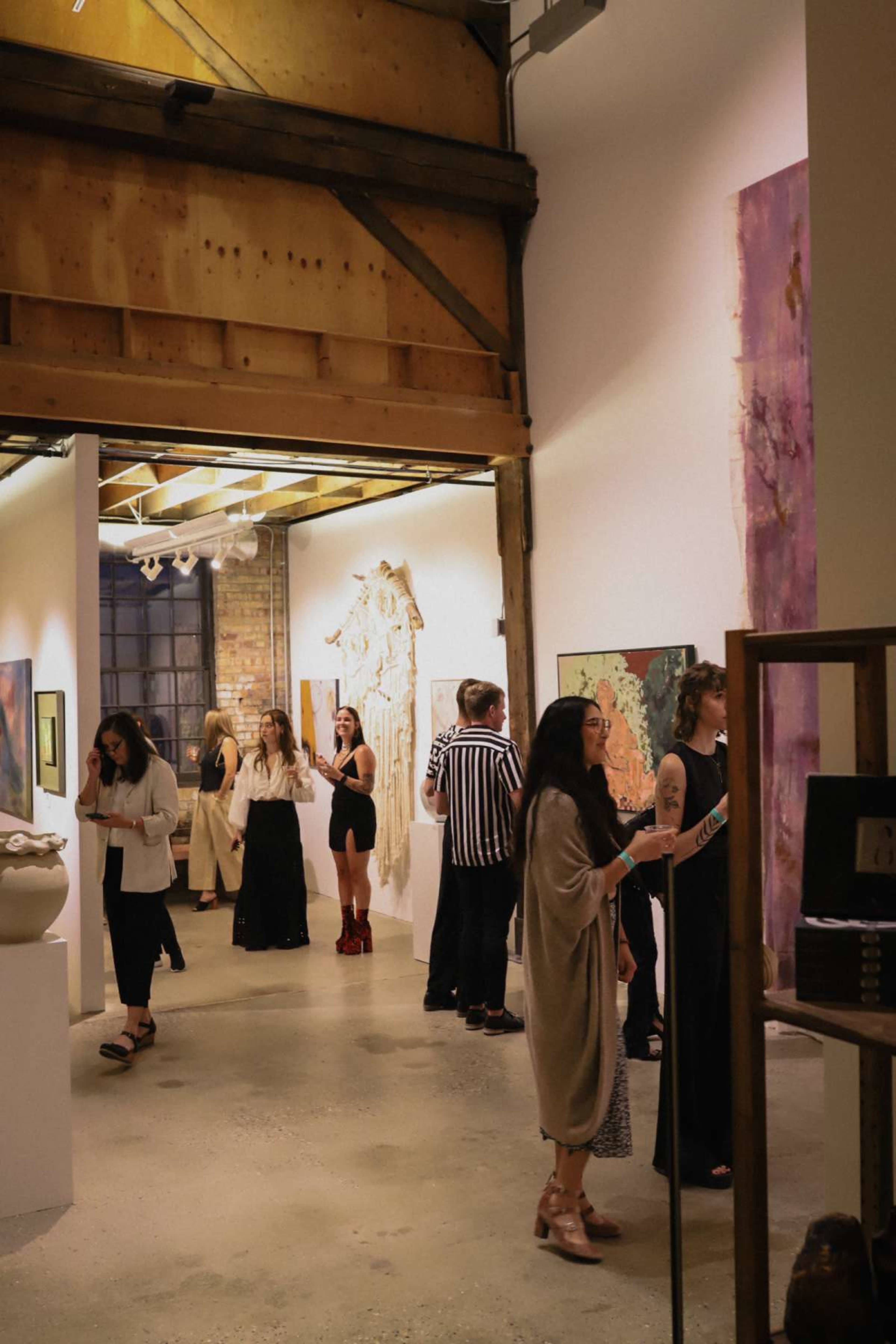 The image shows a gallery filled with people observing various artworks displayed on the walls and pedestals.
