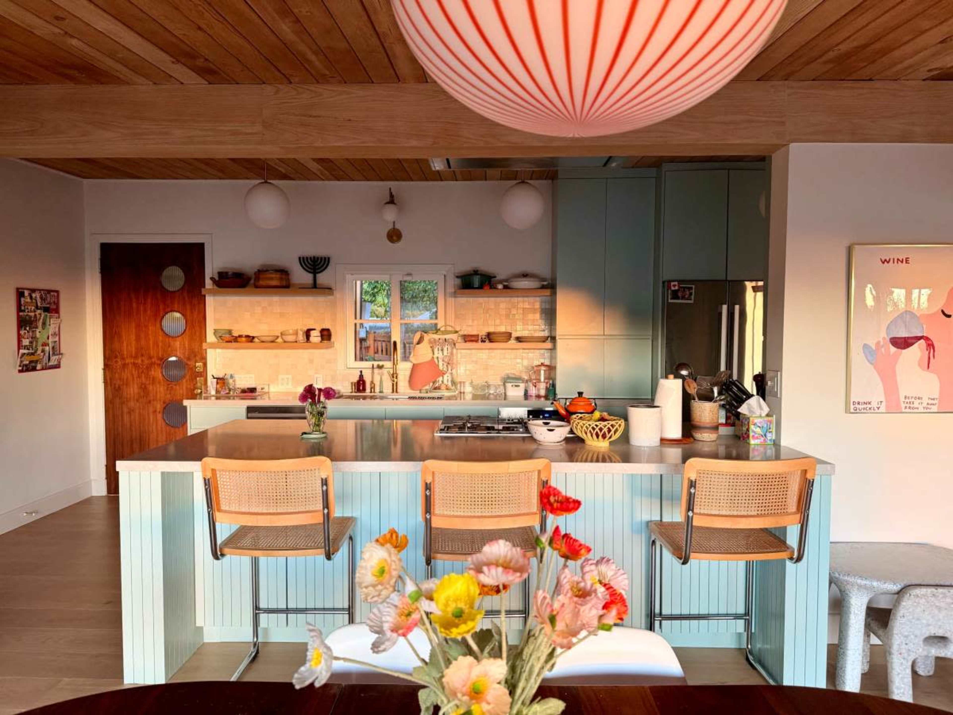 The image shows a modern kitchen with a light blue island, wooden cabinetry, and a dining area featuring a round table with a vase of flowers.
