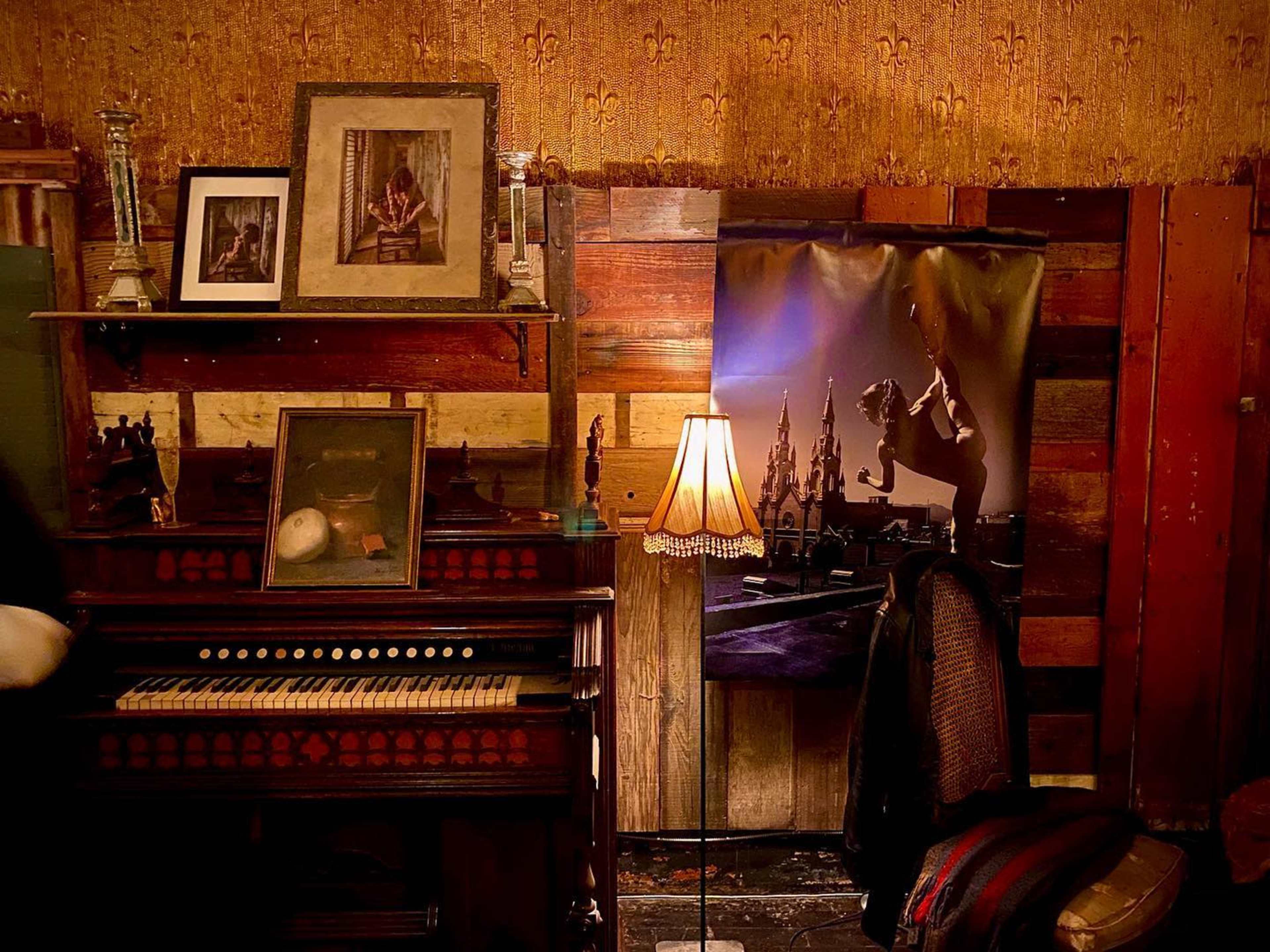 The scene features an antique piano surrounded by framed pictures on a wooden wall, with a decorative lamp casting light nearby.