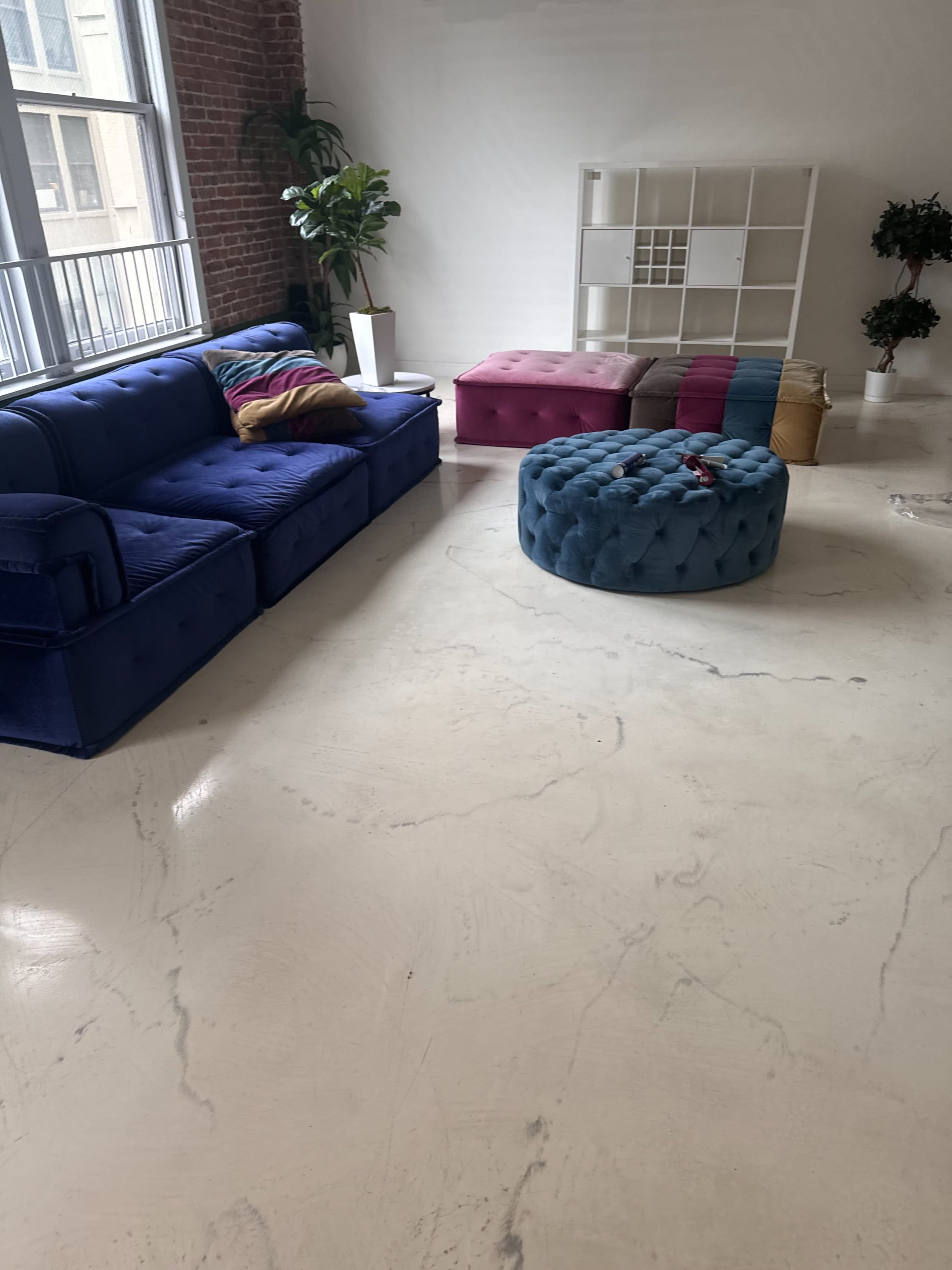 A spacious interior with a blue sofa, a round tufted ottoman, and a storage shelf, all set against a backdrop of large windows and exposed brick walls.