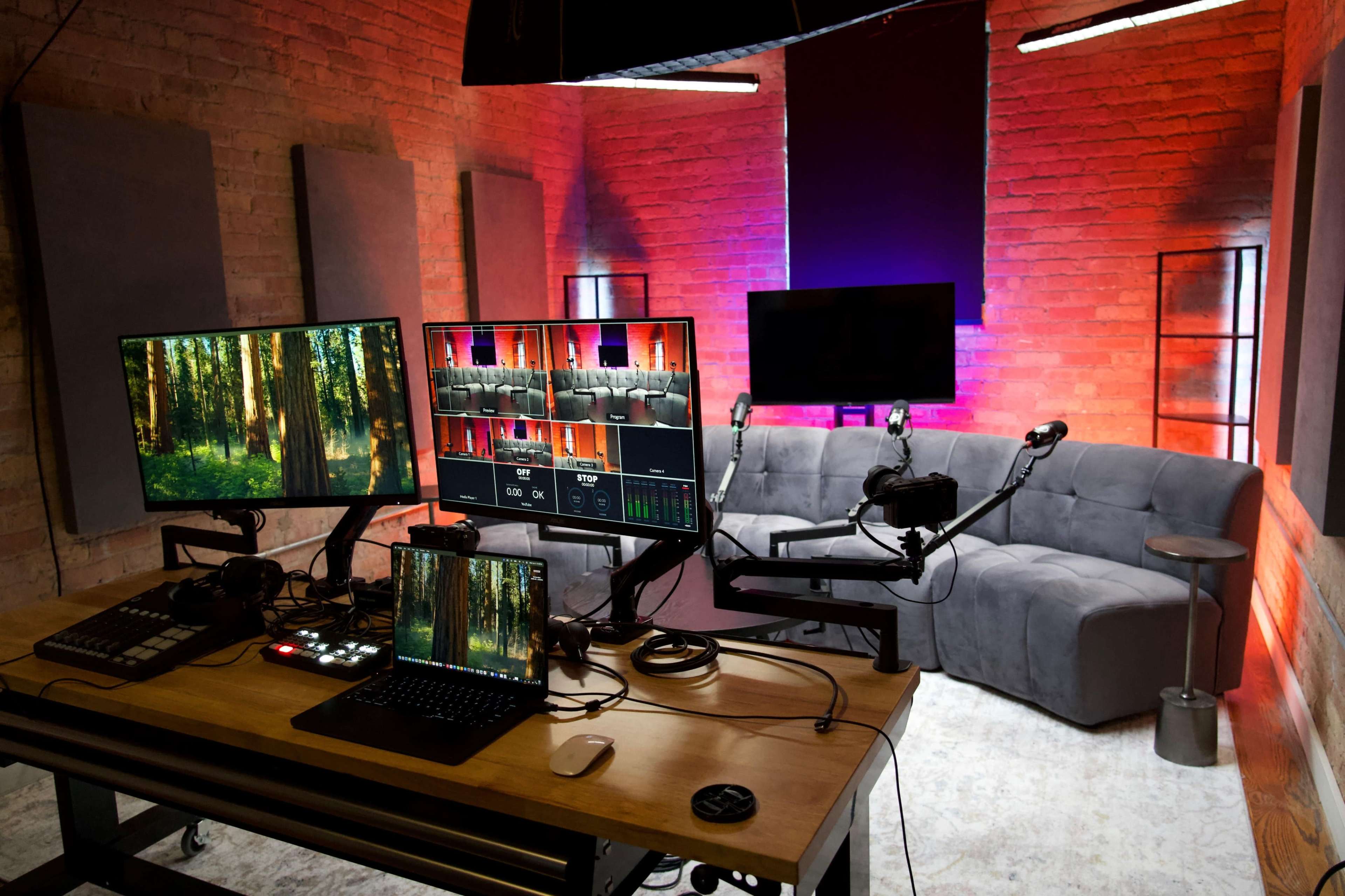 The image shows a modern studio setup with multiple monitors, a laptop, and microphones arranged on a large table, alongside a gray sofa against a brick wall illuminated with colored lights.