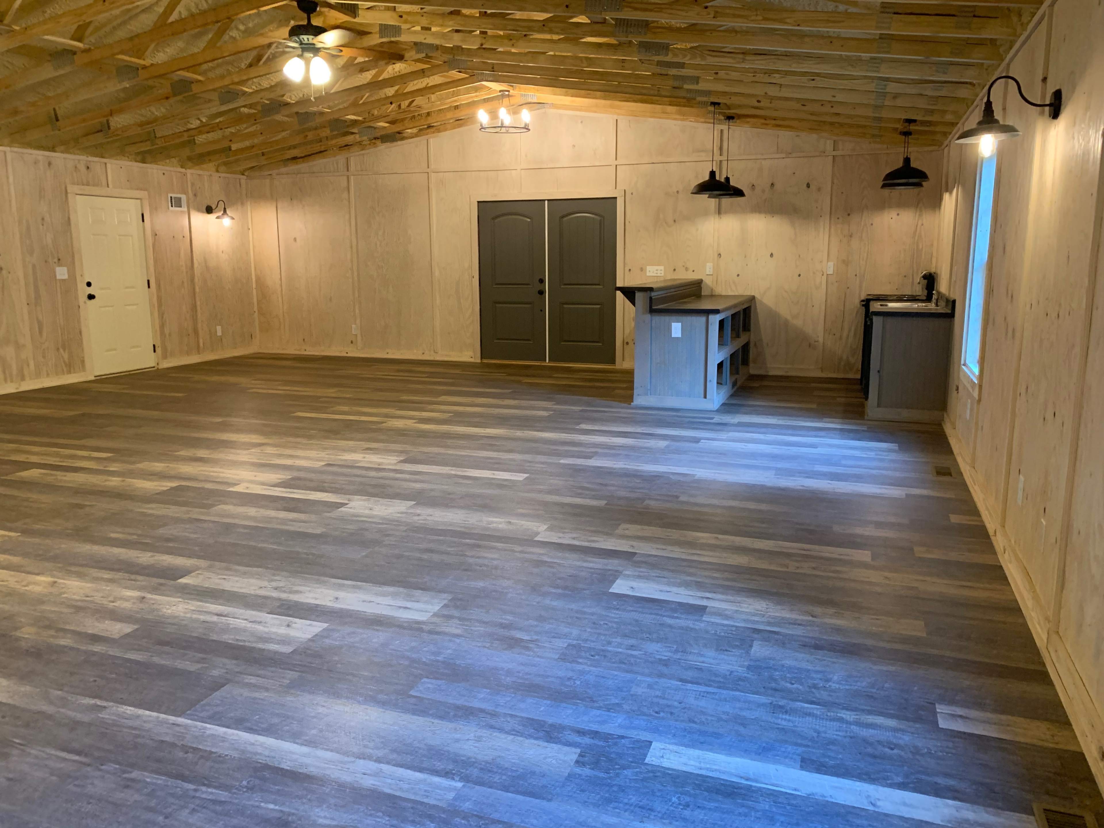 The image shows a spacious, unfinished room with wood-paneled walls, a high ceiling, and a wooden floor, featuring two doors and a small kitchenette in the corner.