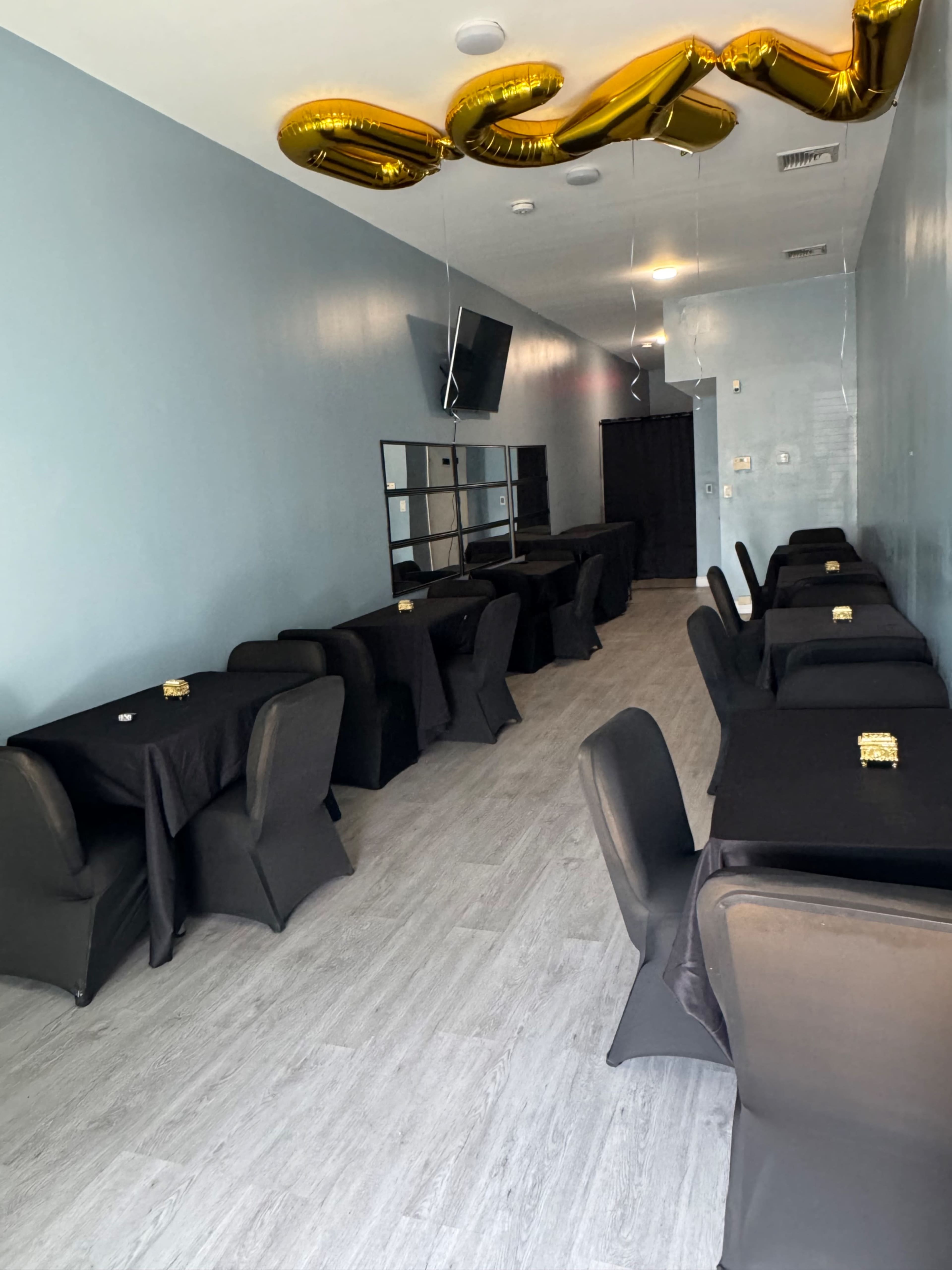 The image shows a spacious, empty dining area with black tablecloths on twelve tables arranged in rows along a light blue wall, adorned with golden balloons on the ceiling.