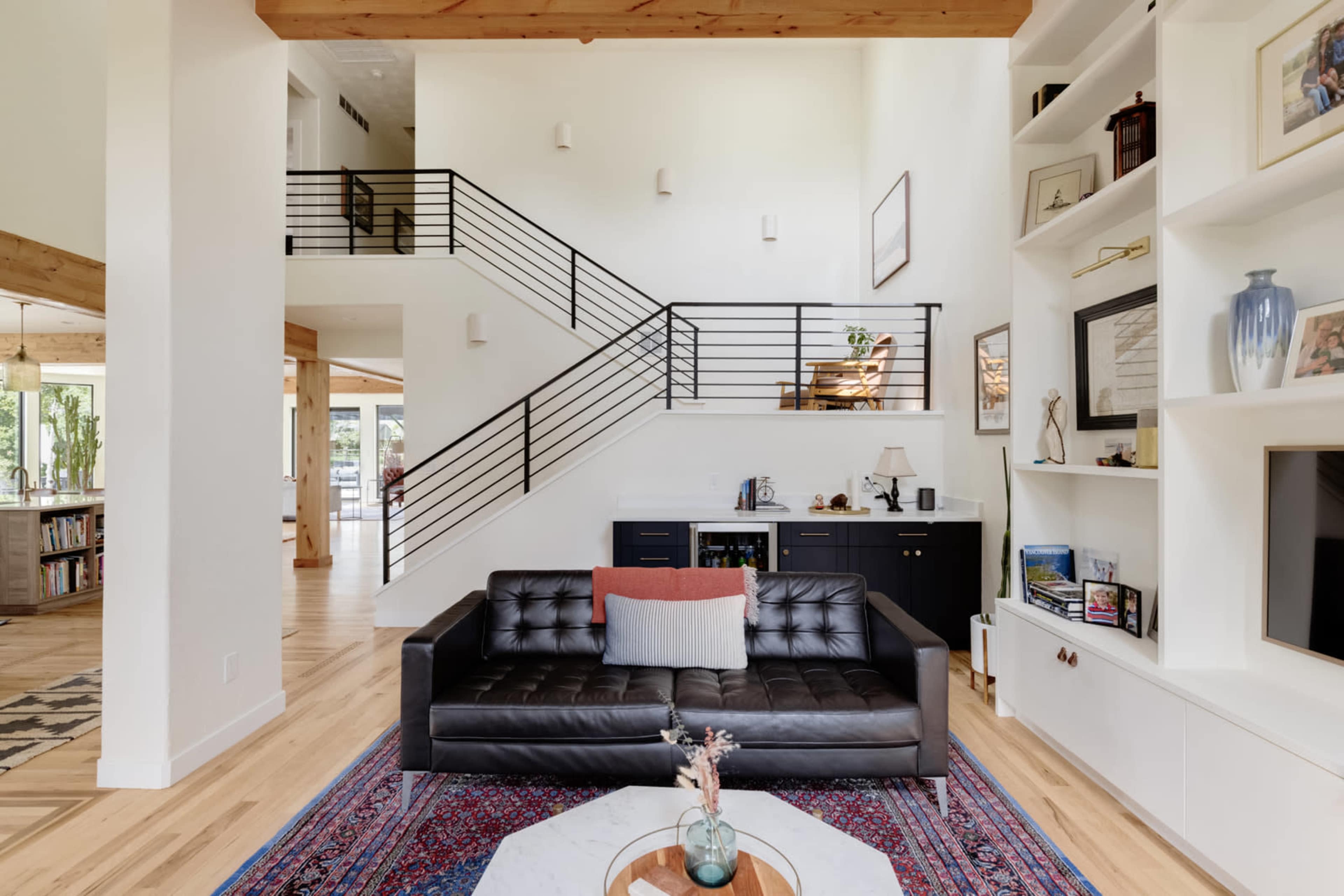 The image shows a modern living room featuring a black leather sofa, a patterned rug, a white coffee table with a flower vase, and a staircase with metal railings in the background.