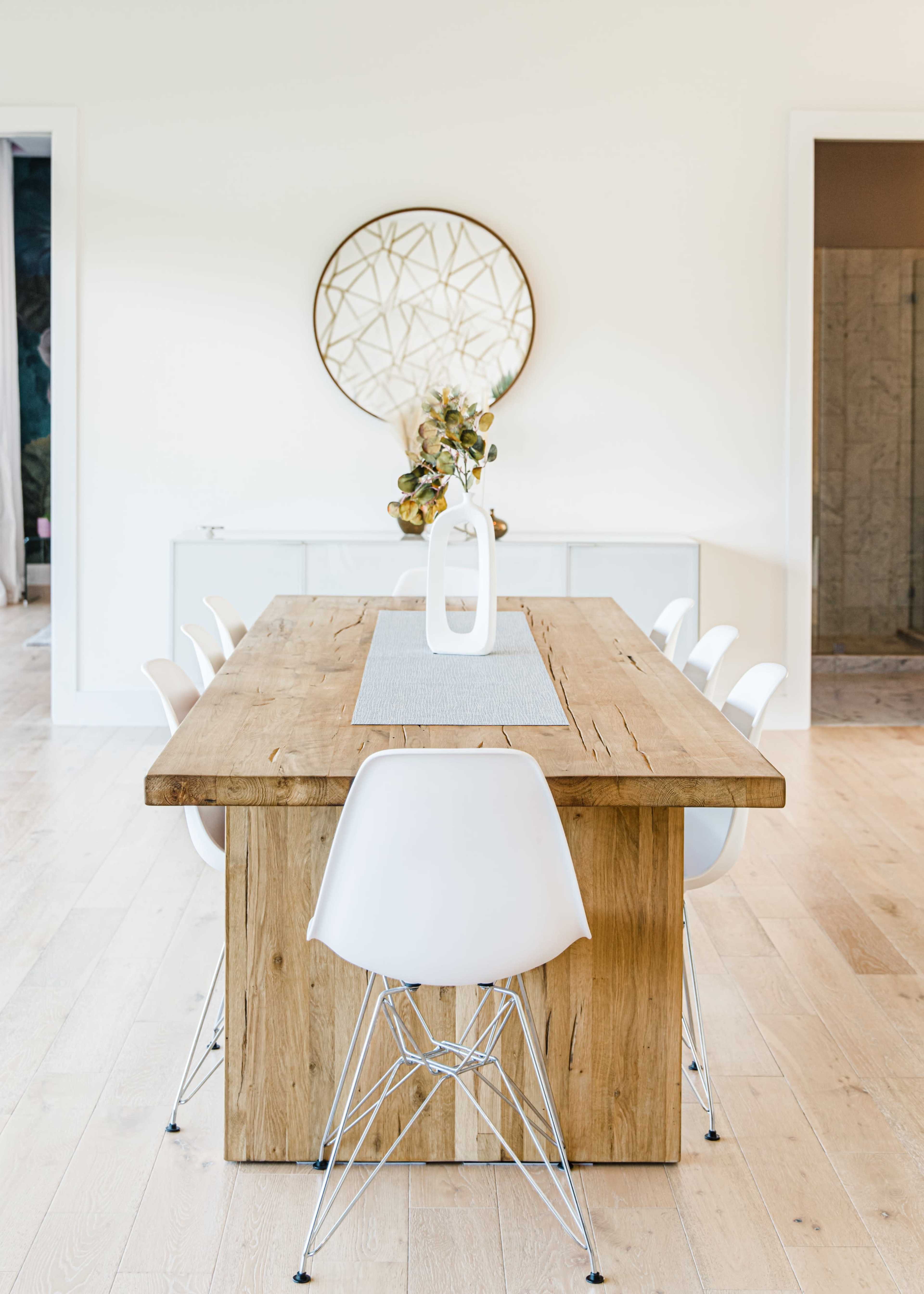 A wooden dining table with a central vase and a runner is surrounded by white chairs in a brightly lit room.