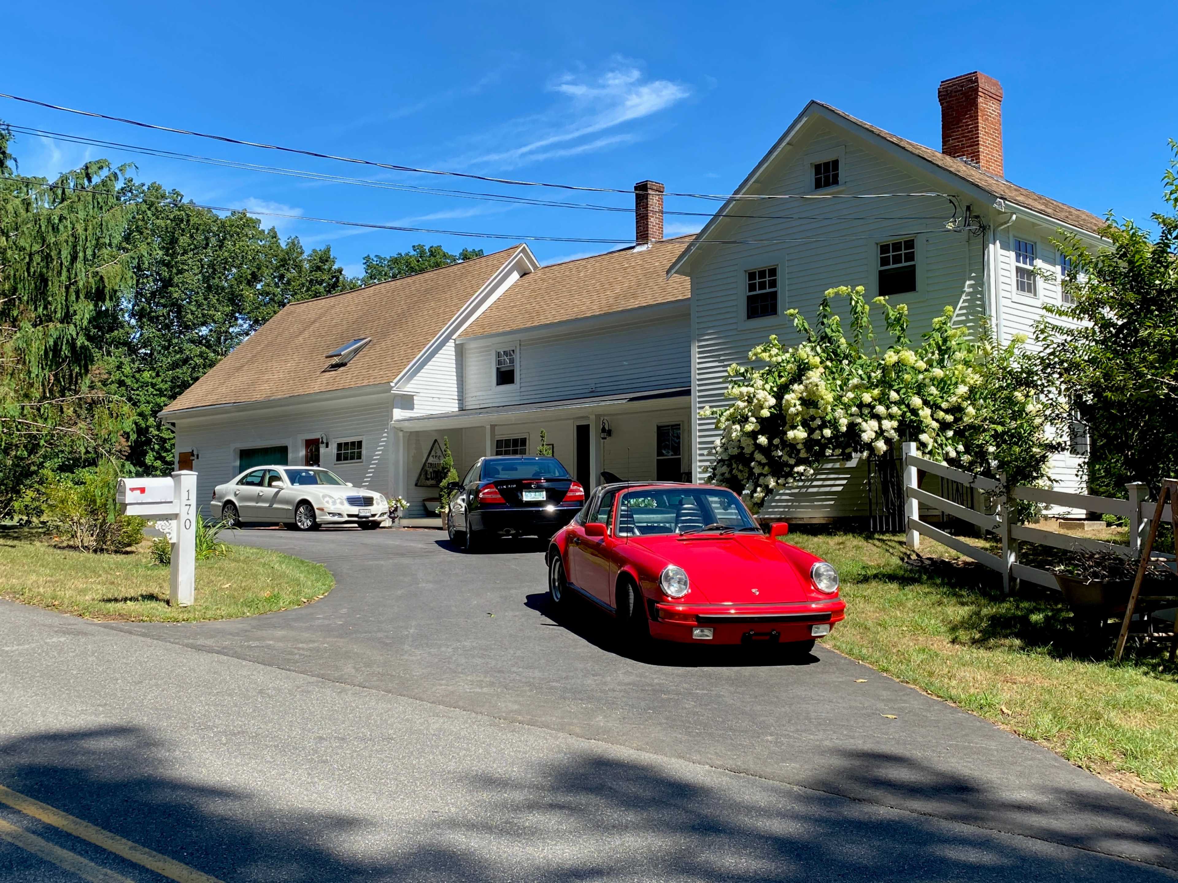 A red sports car and two other vehicles are parked in front of a large white house surrounded by greenery.