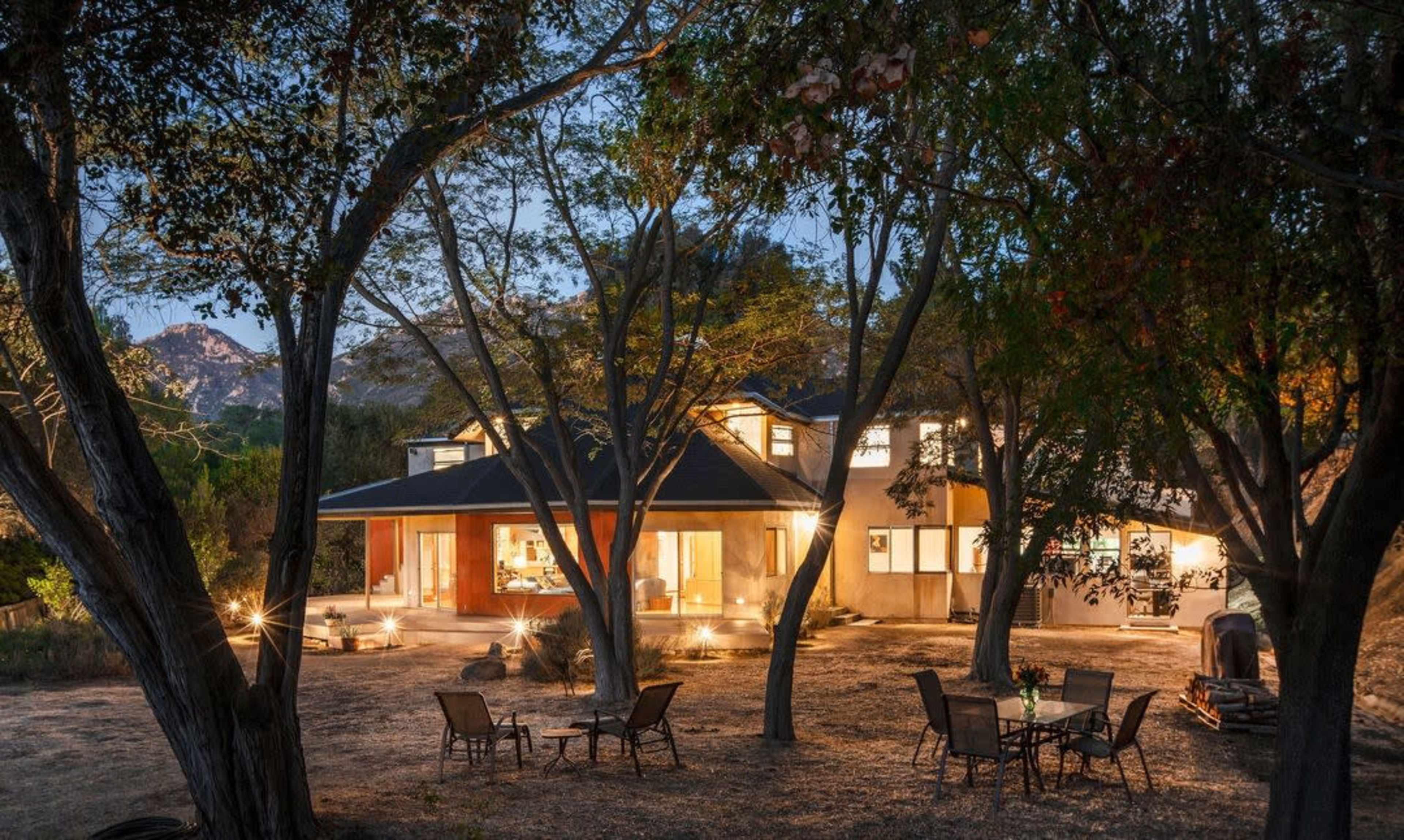 A modern house is surrounded by trees, with outdoor seating arranged in a clear area, illuminated softly by outdoor lights at dusk.