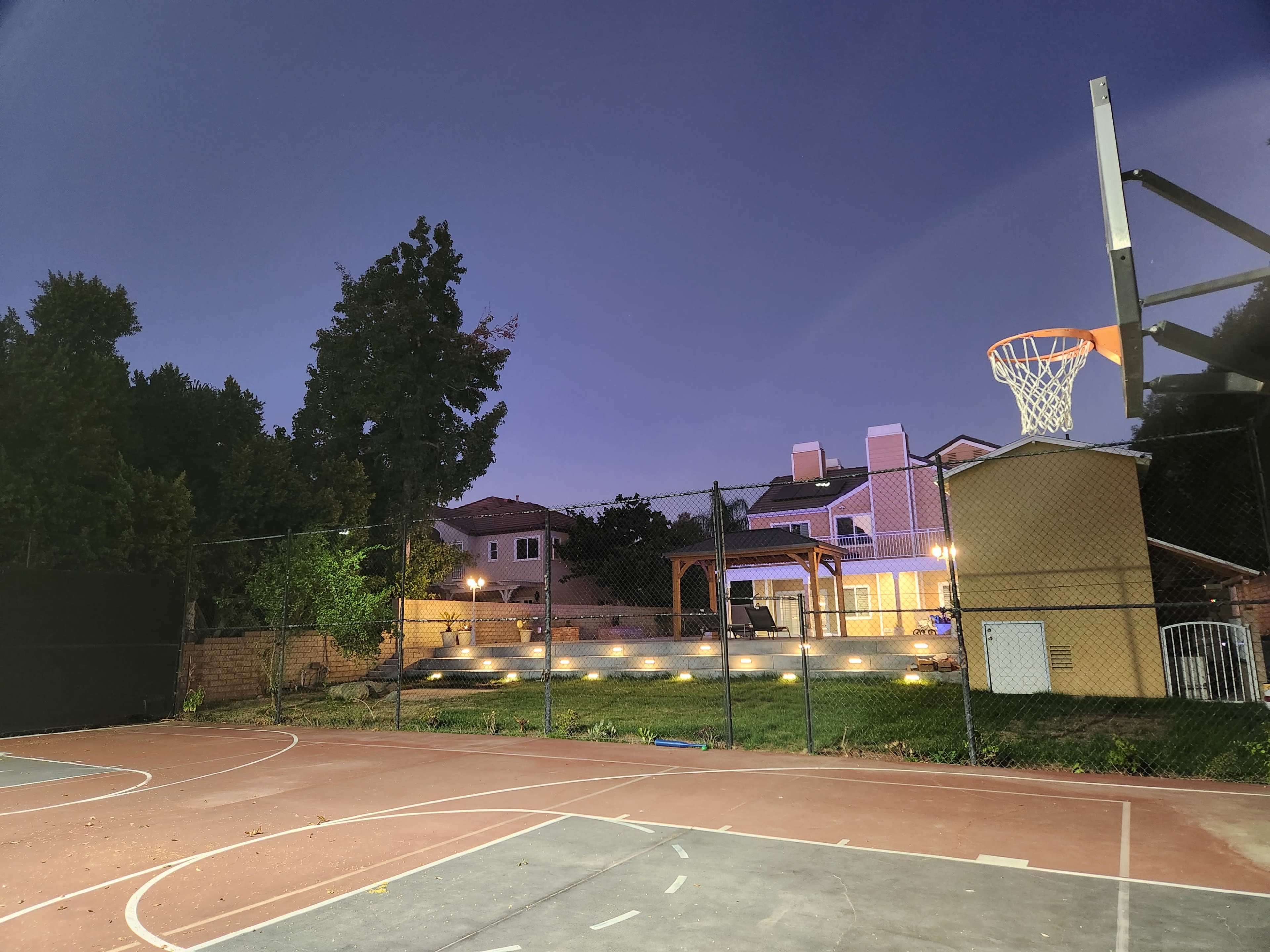The image shows a basketball court illuminated by evening lights, with a residential house and landscaped area visible in the background.