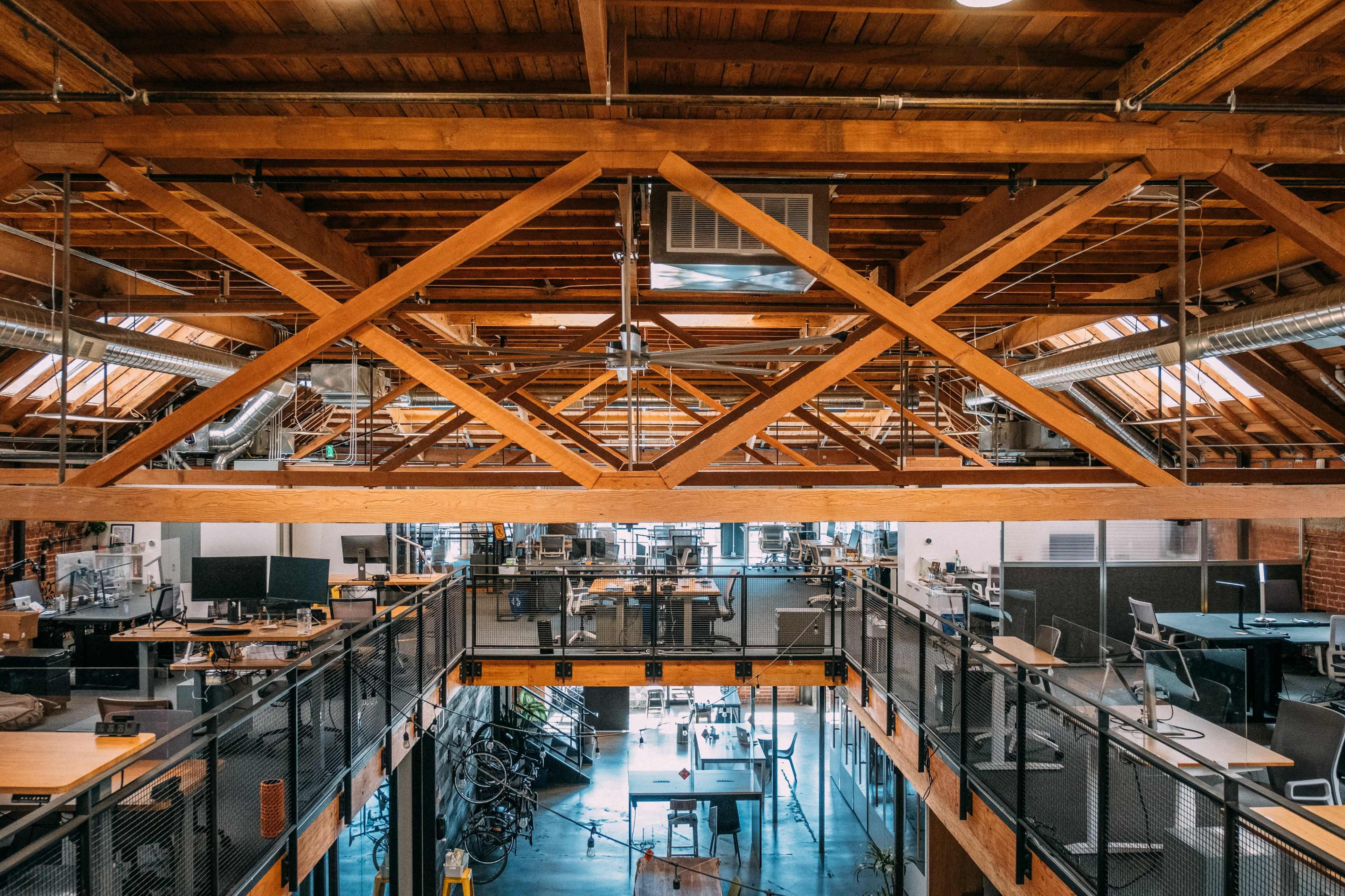 An open-concept office space with wooden beams, multiple workstations, and large windows that allow natural light to enter.