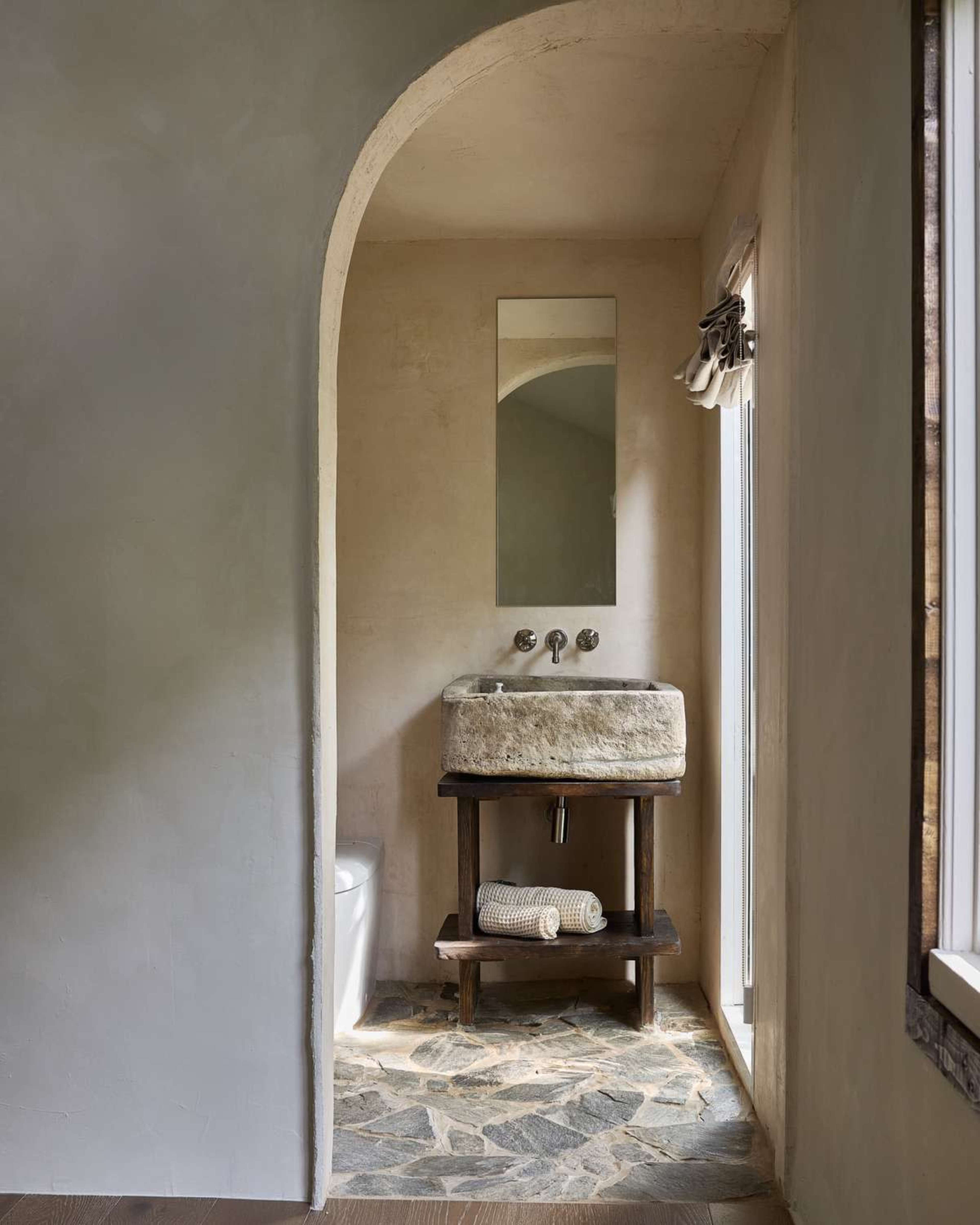 The image shows a minimalist bathroom featuring a stone sink on a wooden stand, a large mirror above the sink, and a window with natural light streaming in.