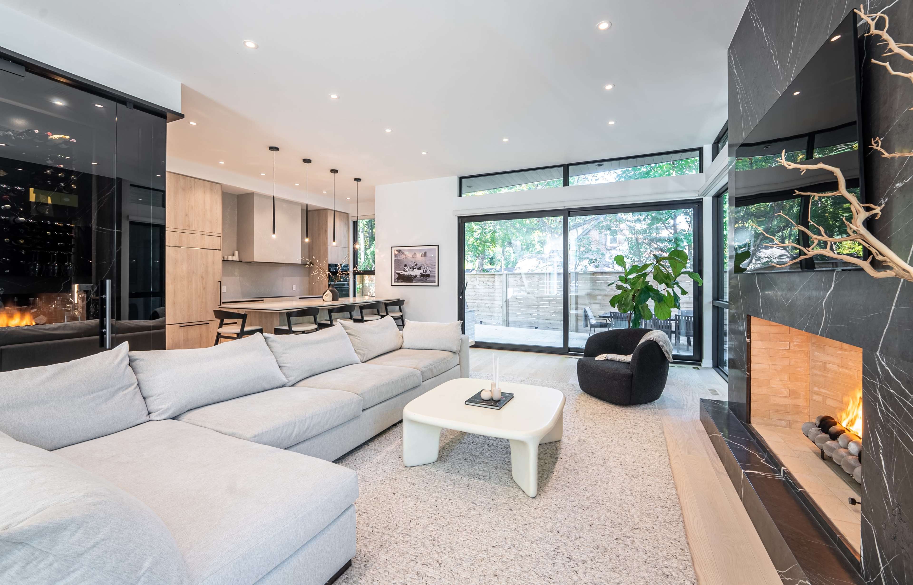 The image shows a modern living room with a large sectional sofa, a coffee table, a stylish fireplace, and windows that provide natural light and views of greenery outside.