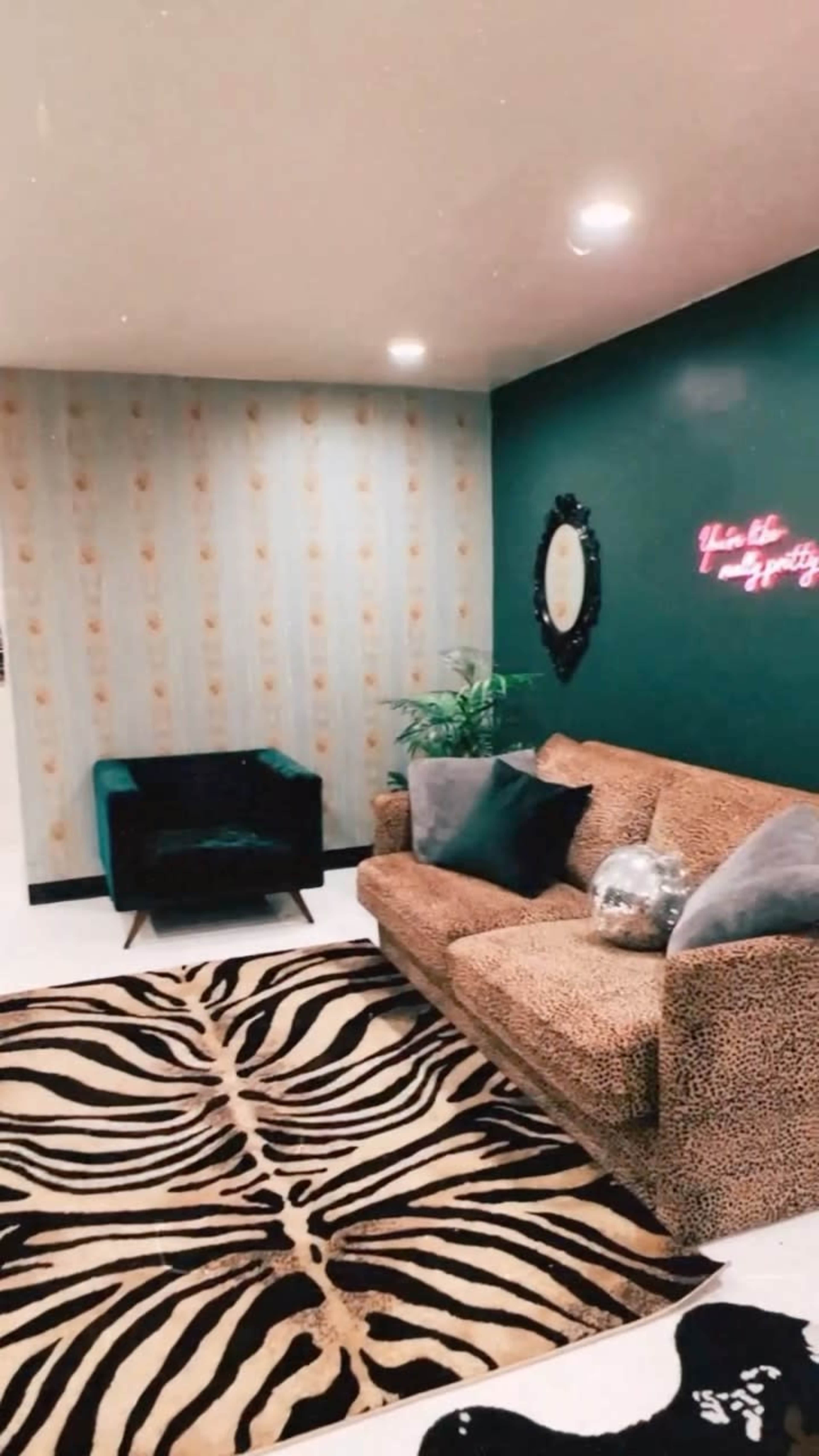 A cozy living room with a leopard print sofa, a black armchair, a zebra-patterned rug, and a wall featuring a decorative mirror and neon artwork.