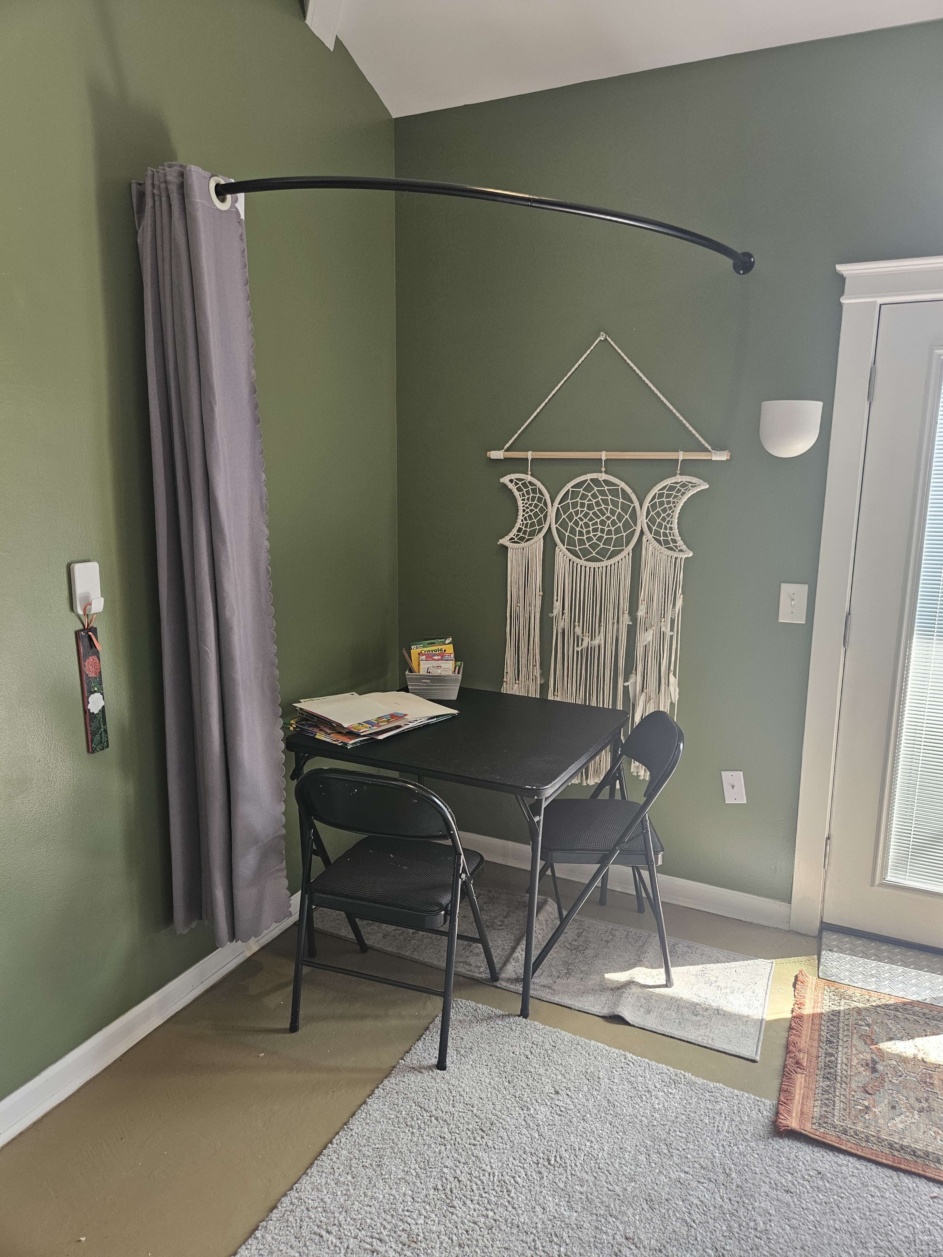 The image shows a small corner of a room with a black folding table and two chairs, draped with a gray curtain and featuring a dreamcatcher hanging on the wall.