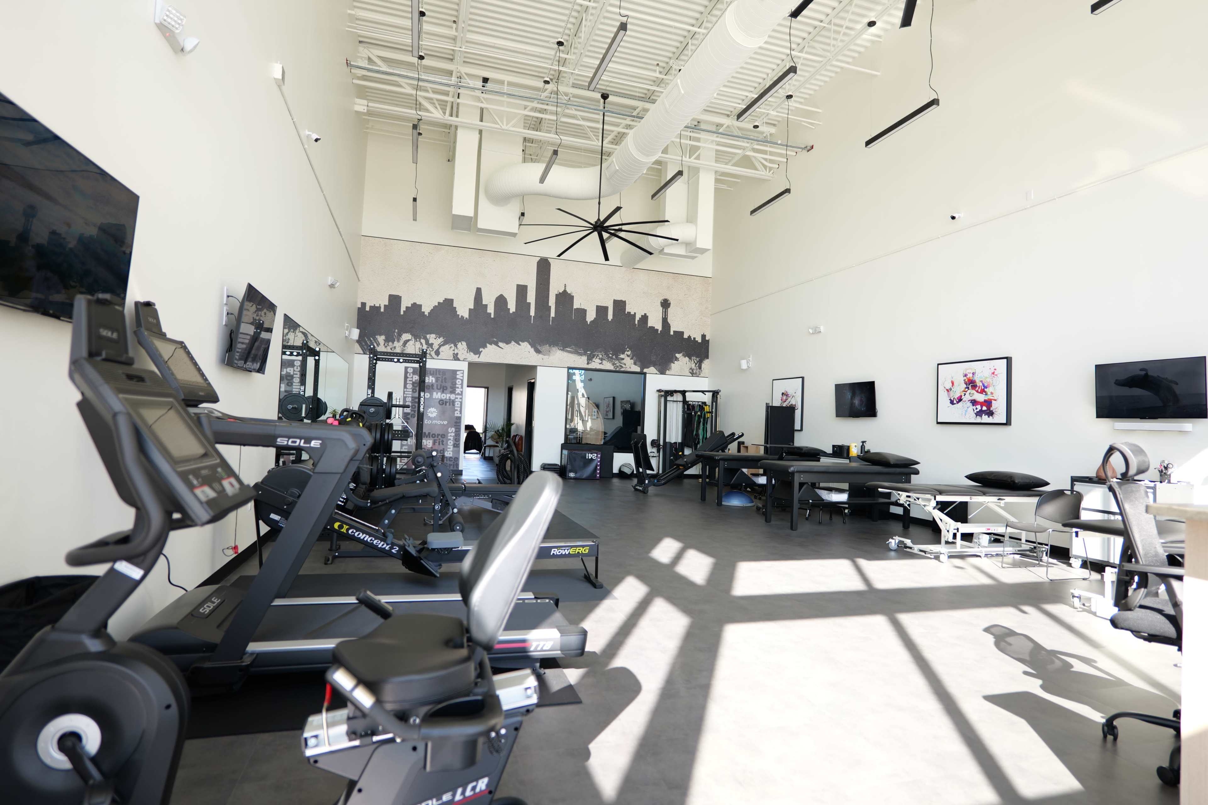 The image shows a modern fitness facility equipped with exercise machines, treatment tables, and large windows allowing ample natural light.