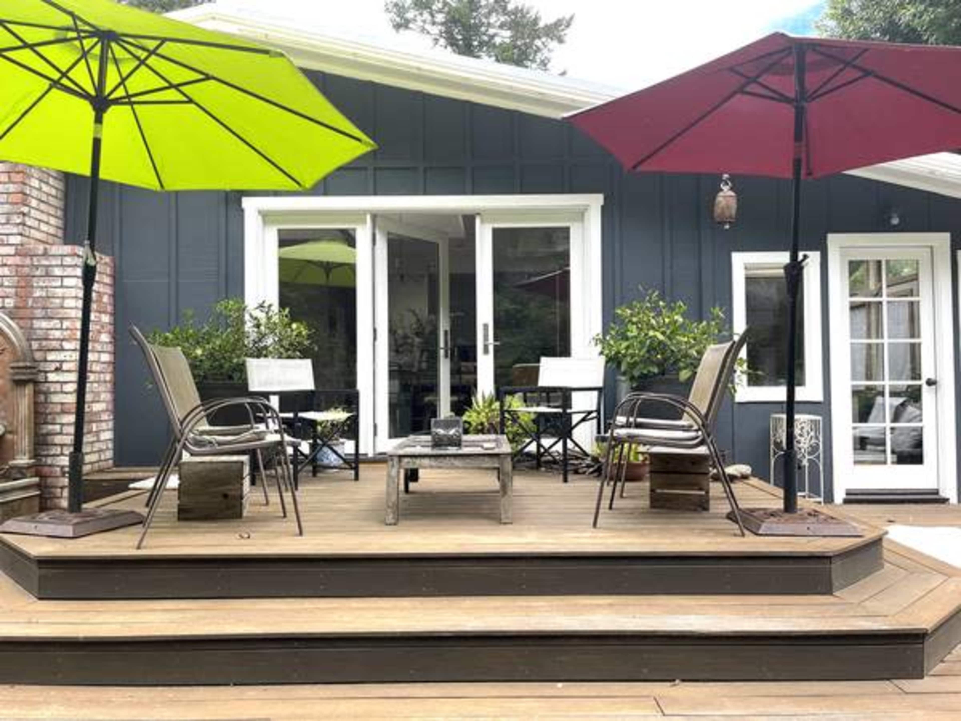 A wooden deck with two chairs and a table, surrounded by colorful umbrellas, leading to a house with large windows.