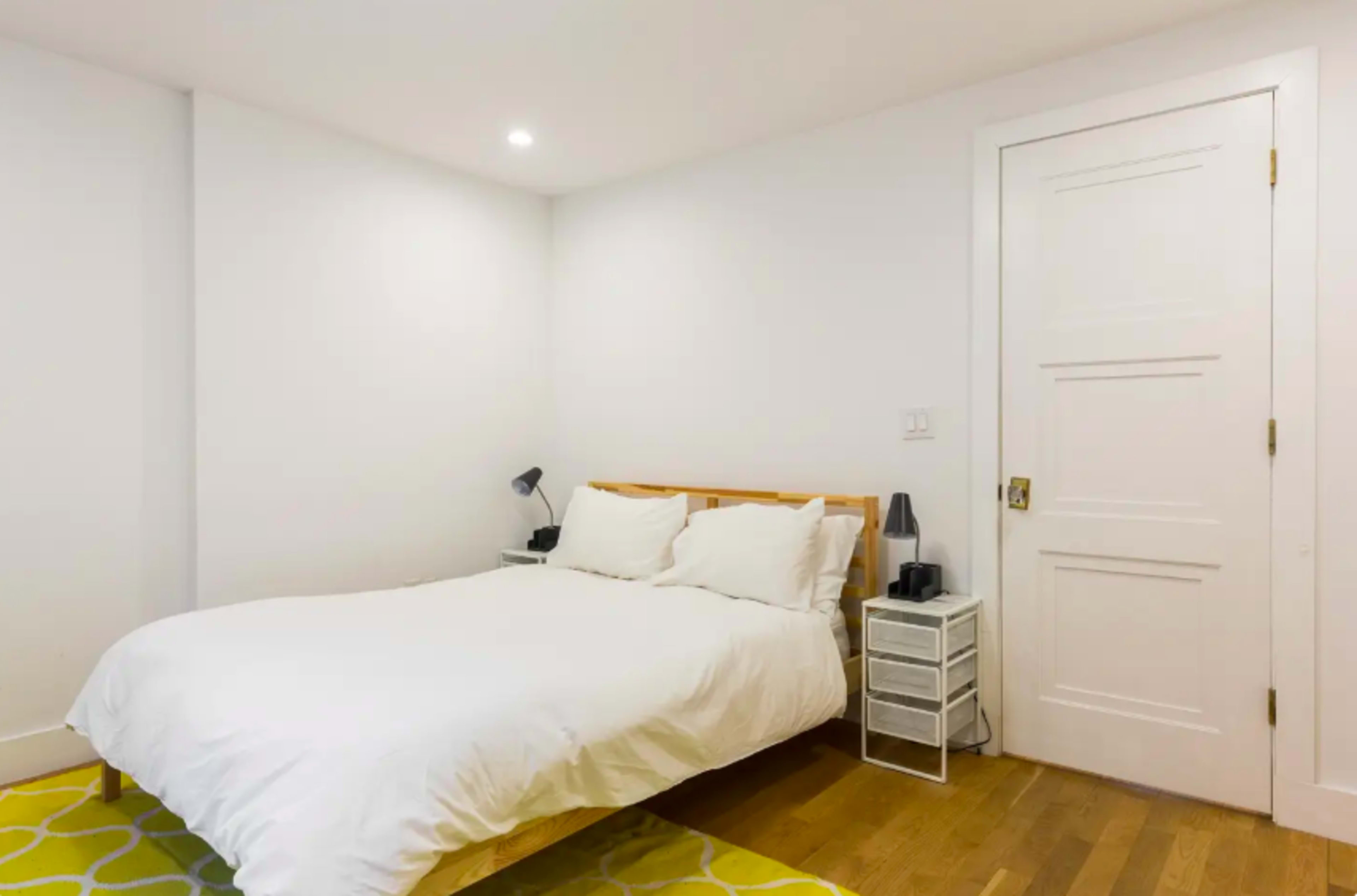 A simple bedroom with a wooden bed, white bedding, a nightstand with lamps, and a door on the opposite wall.