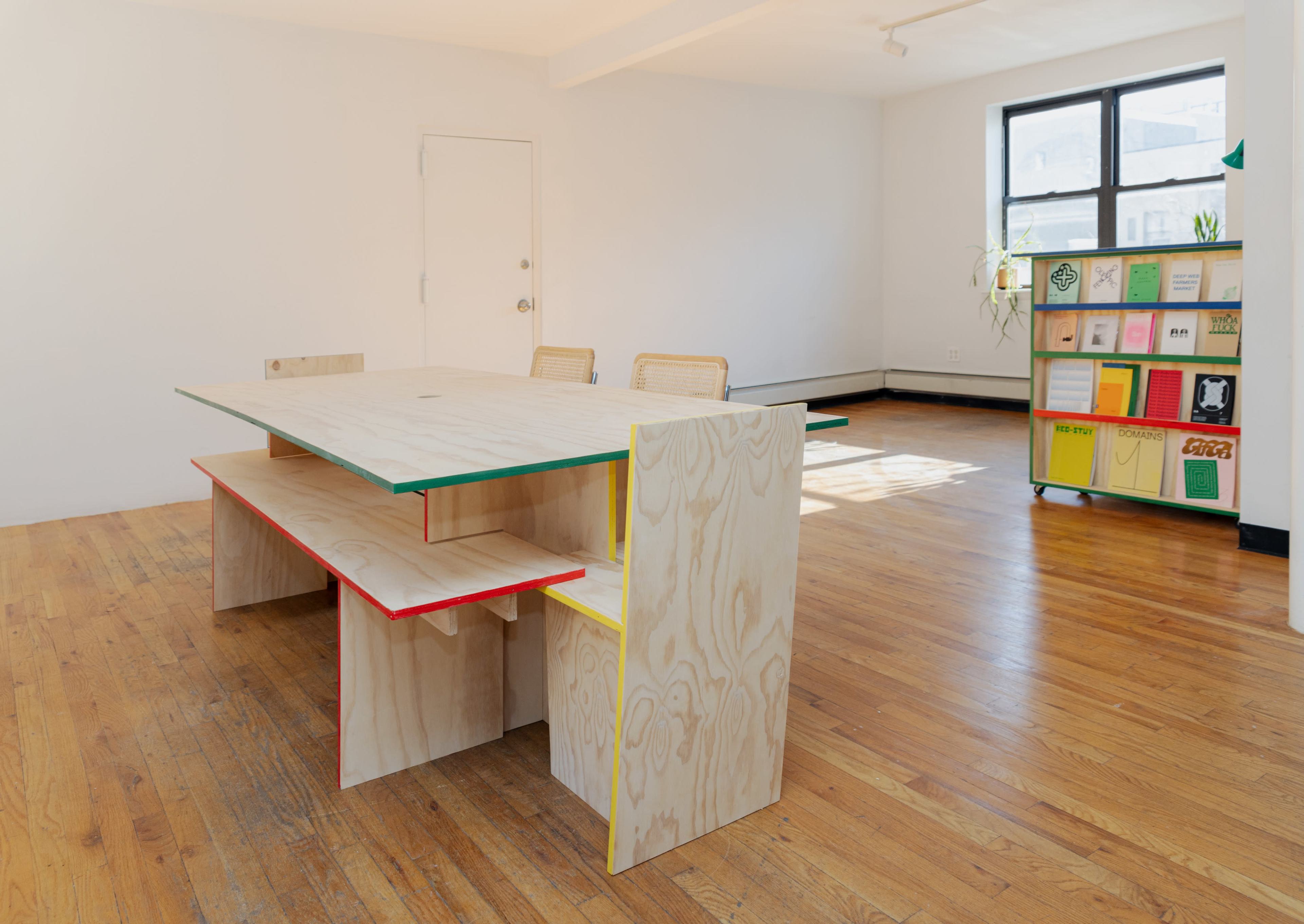 A large, minimalist wooden table with colorful accents is positioned in a well-lit room featuring a bookshelf filled with various materials.